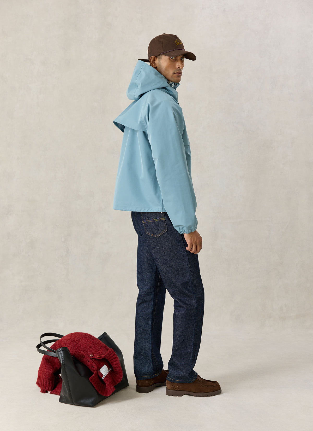 A model wearing the Percival light blue Hanson boxy rain jacket with dark jeans and a brown cap, with a bag and red knit.