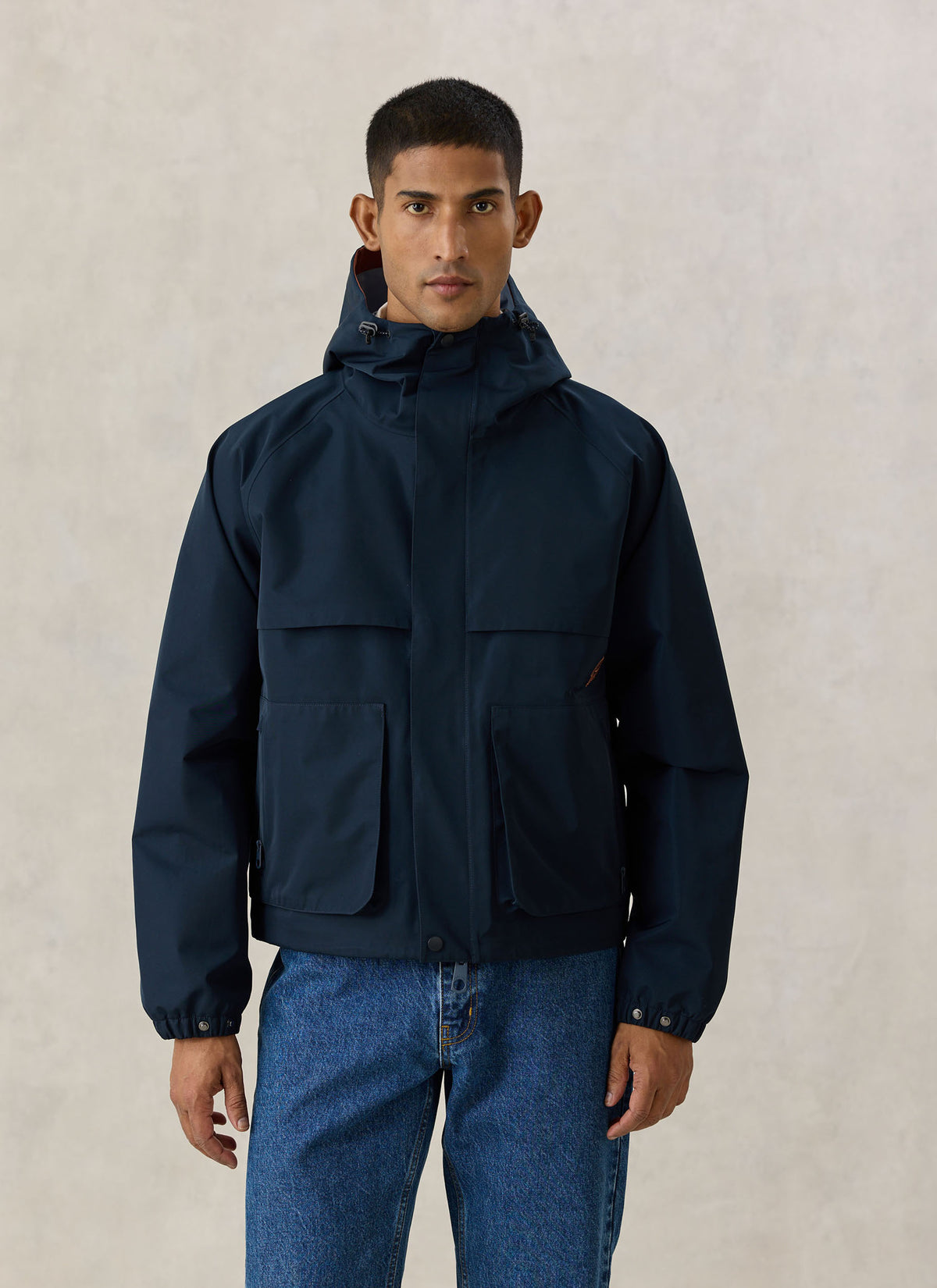 A model wearing the Percival navy blue Hanson boxy waterproof rain jacket with a hood and large front pockets.
