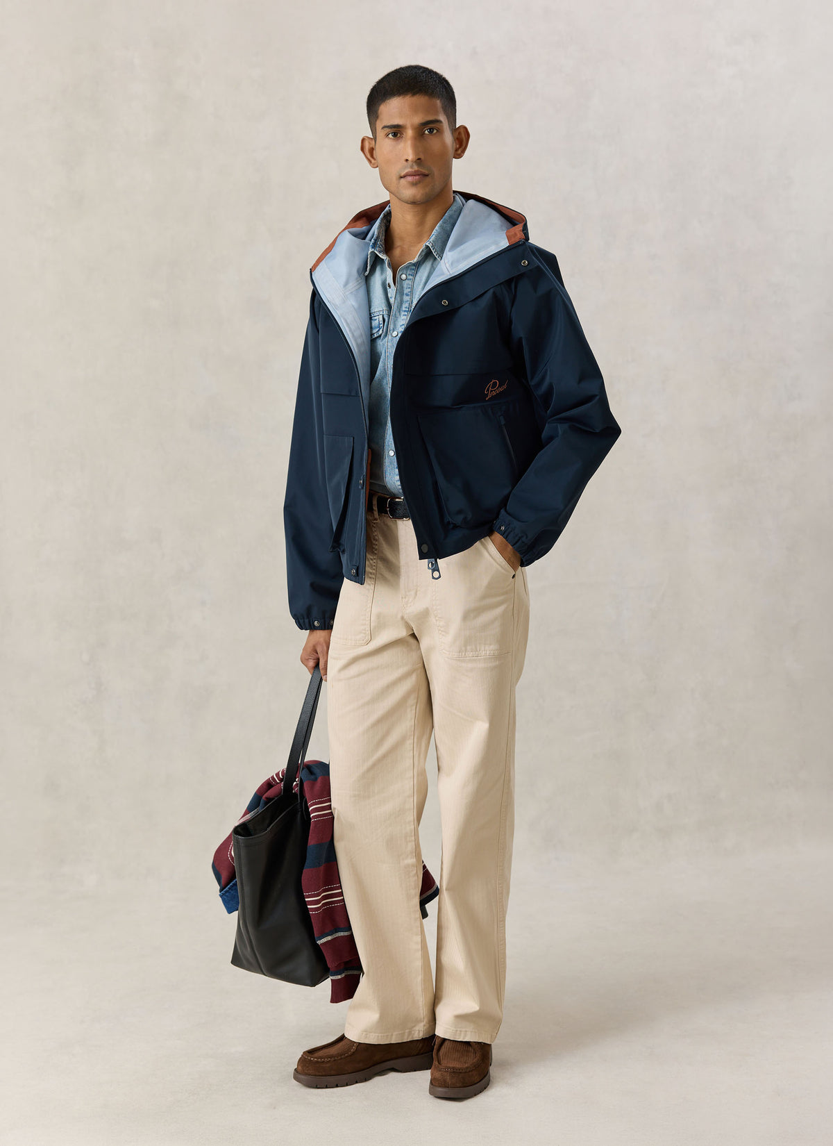 A model wearing the navy Percival Hanson waterproof boxy rain jacket over a light blue denim shirt and beige trousers.