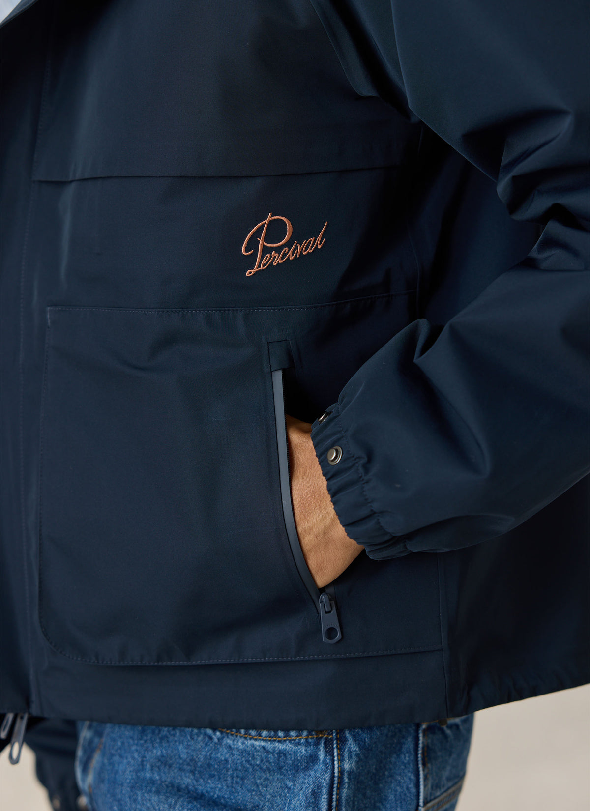 A close-up of the navy waterproof Percival rain jacket showing the embroidered logo and zippered front pocket.
