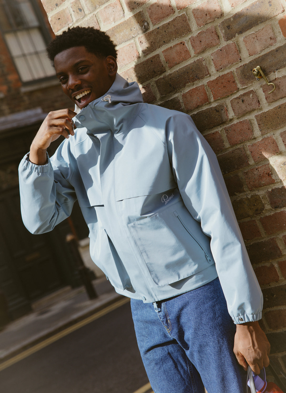 A smiling model wearing the light blue Percival Hanson boxy waterproof rain jacket outdoors against a brick wall.