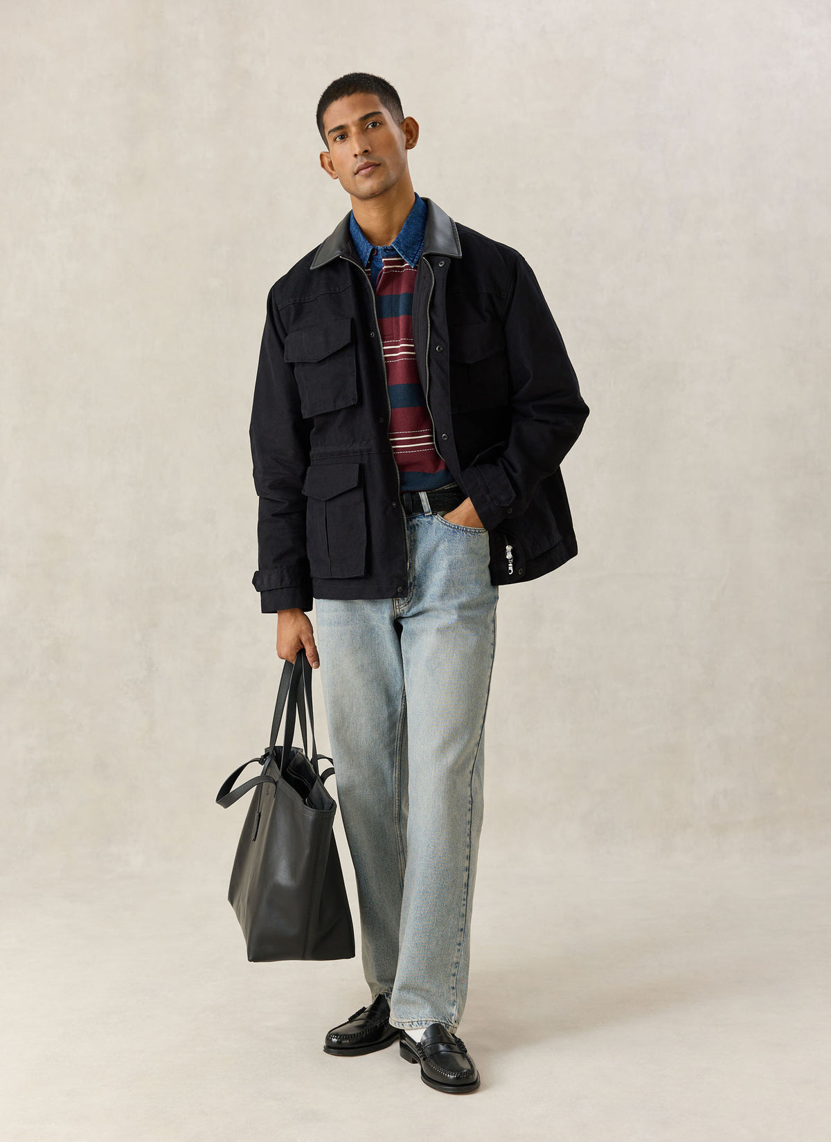 A model wearing the black Percival Harris Field Jacket with a striped knit, light-wash jeans, and a black tote bag.