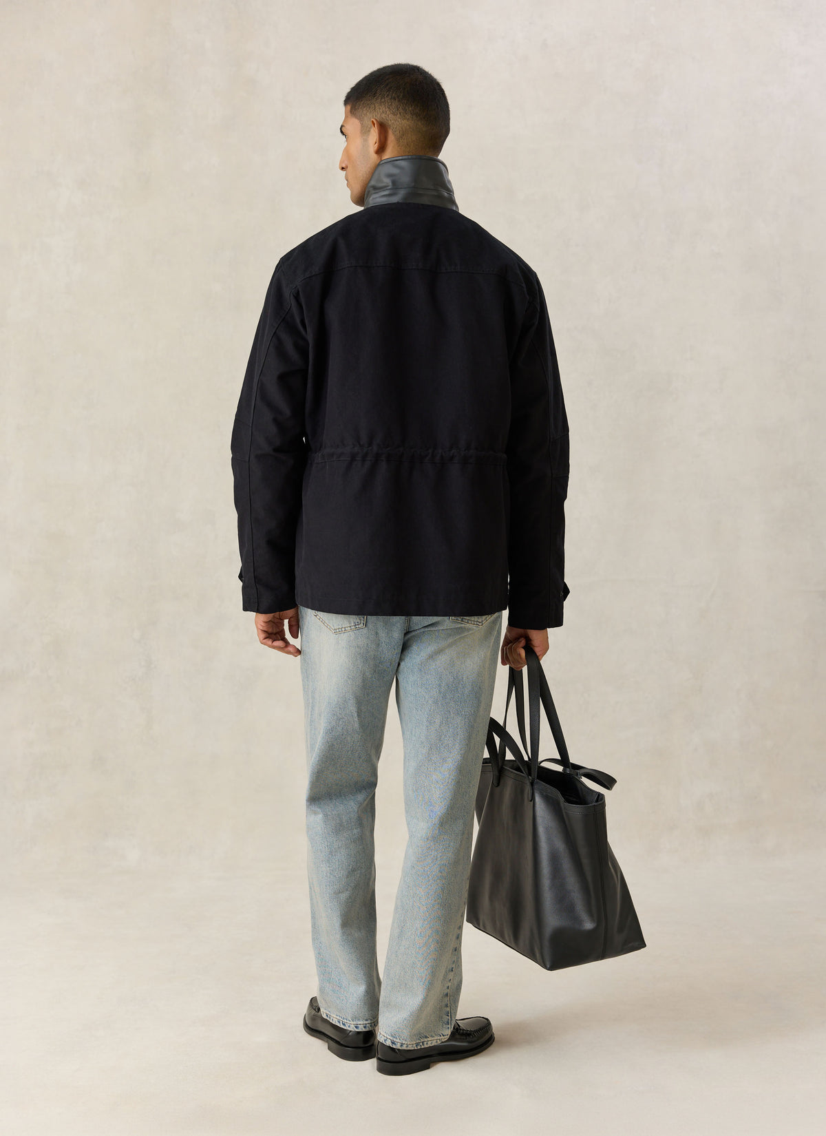 A model from the back wearing the black Percival Harris Field Jacket with a contrasting collar, holding a black tote bag.