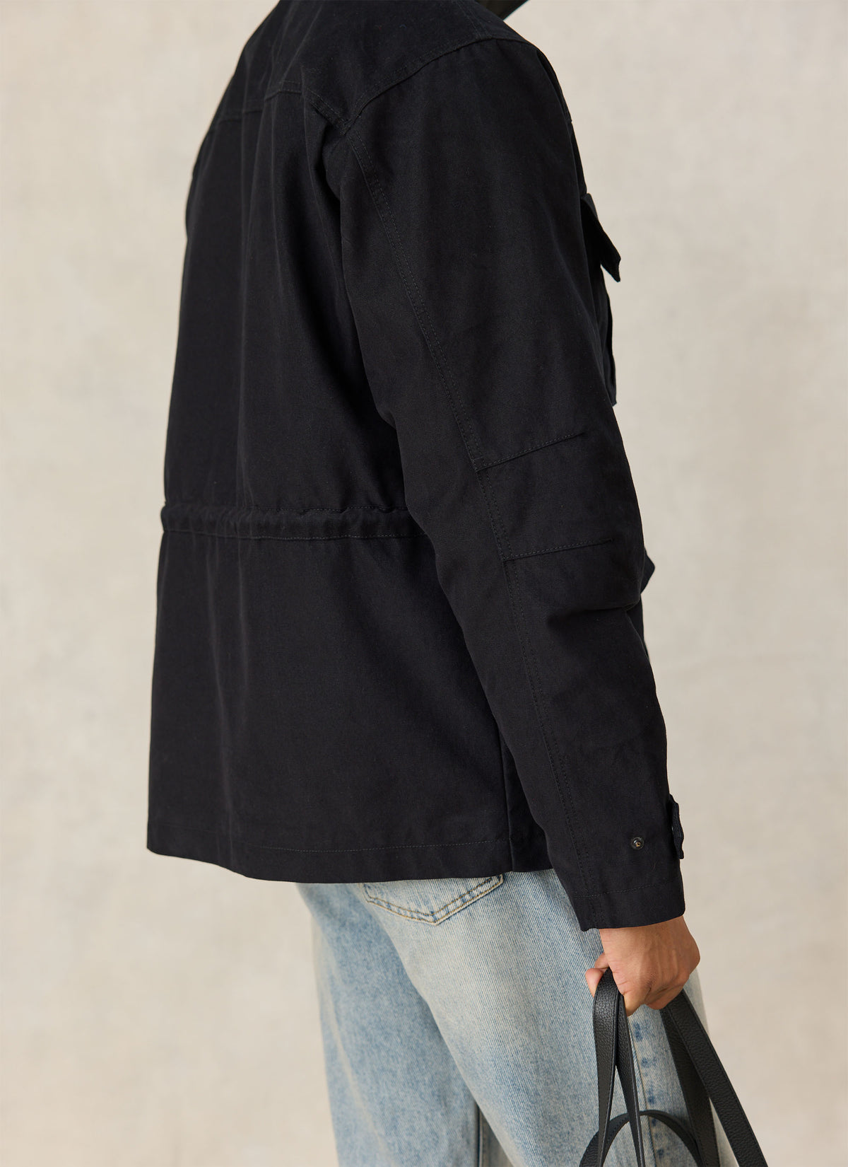 A side view of a model wearing Percival's black Harris Field Jacket paired with light-wash jeans and holding a bag.
