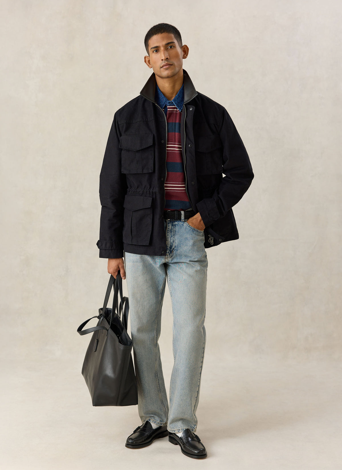 A model wearing the black Percival Harris Field Jacket over a striped polo, blue jeans, and holding a black leather tote bag.