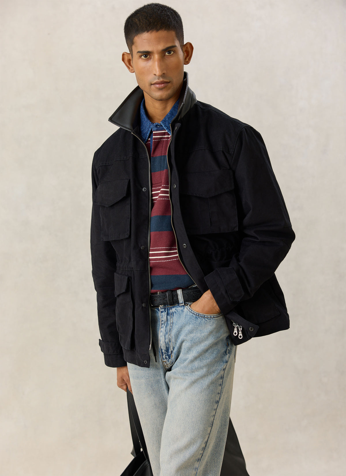 A model wearing the black Percival Harris Field Jacket with four flap pockets over a striped polo shirt and jeans.