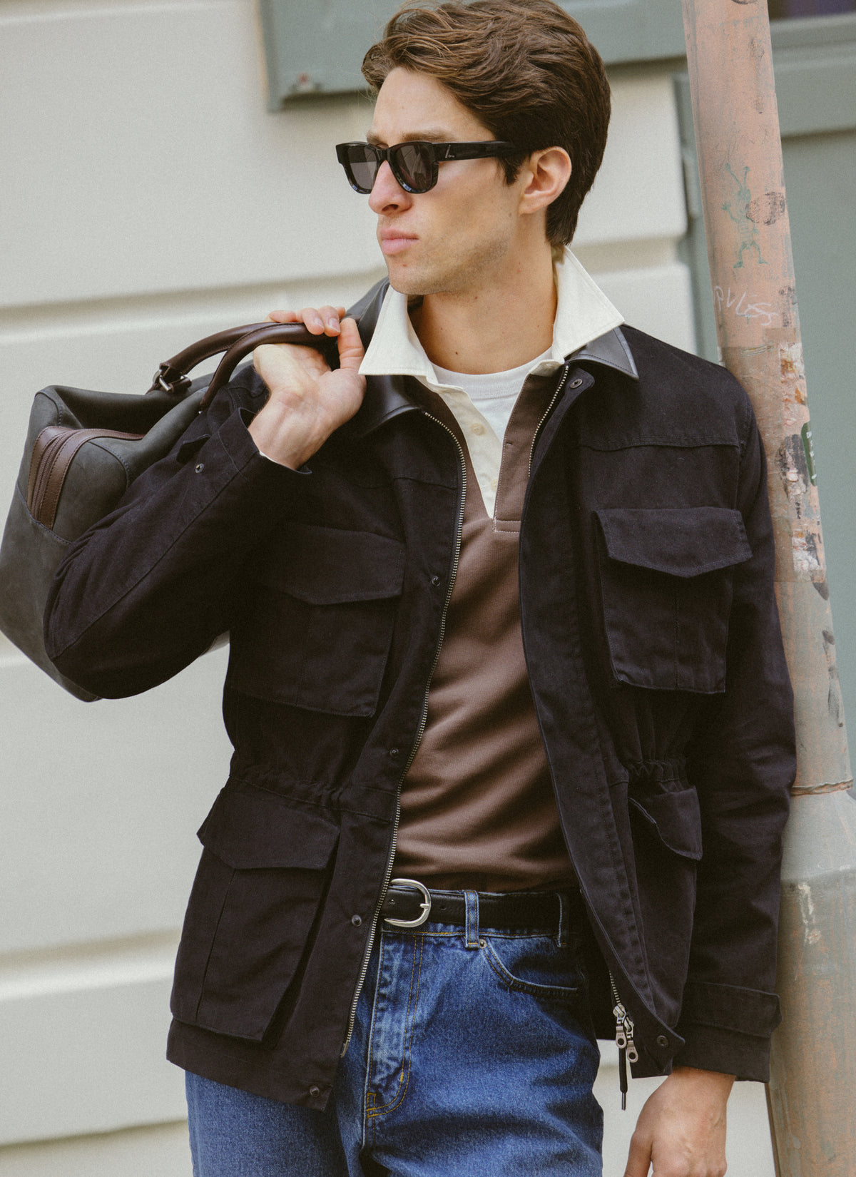 A model wears the black Percival Harris Field Jacket over a brown rugby polo and jeans, holding a bag outdoors.