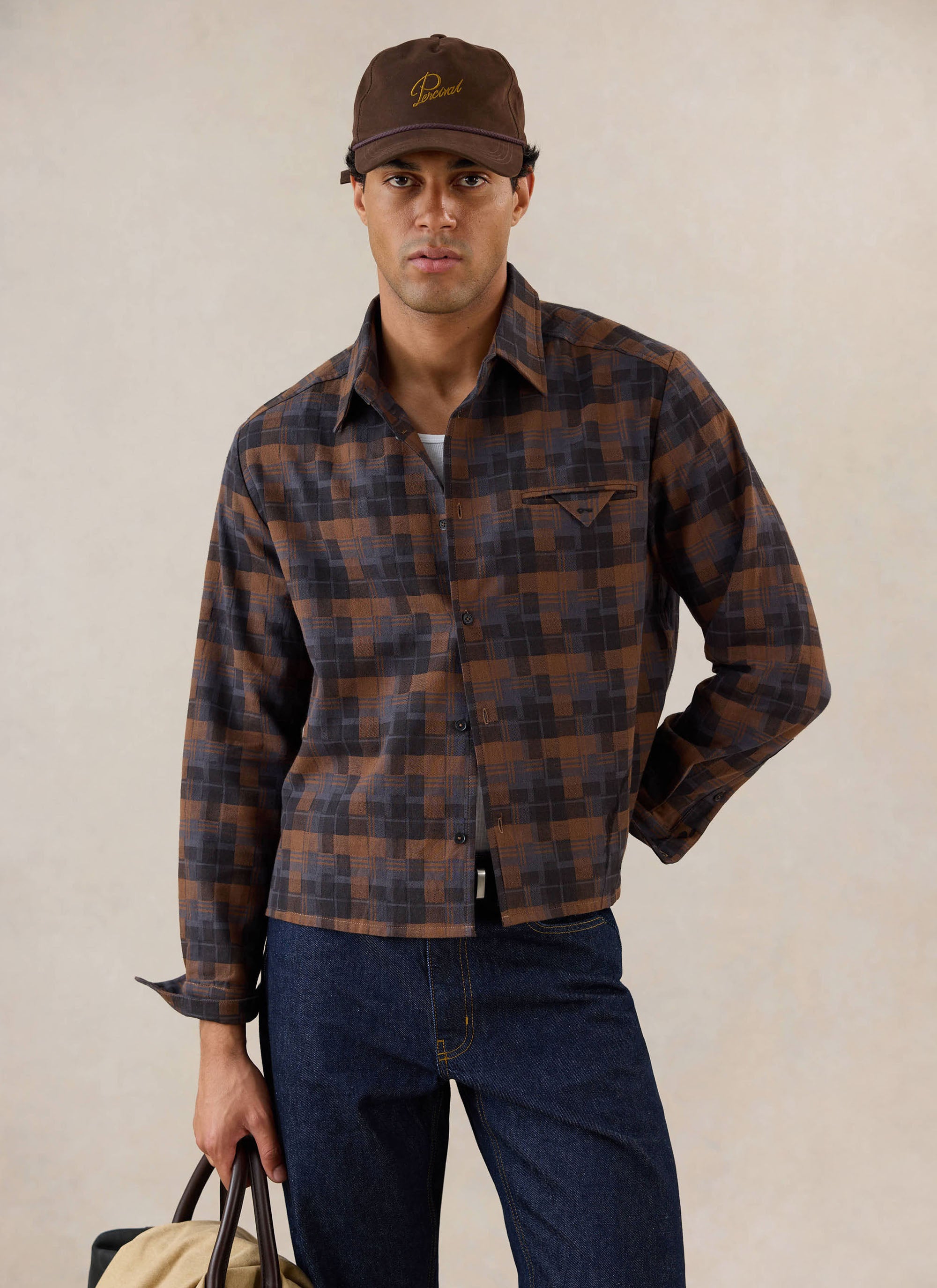 A model wears the brown check Hinsley Boxy Long Sleeve Shirt from Percival, styled with blue jeans and a matching brown cap.