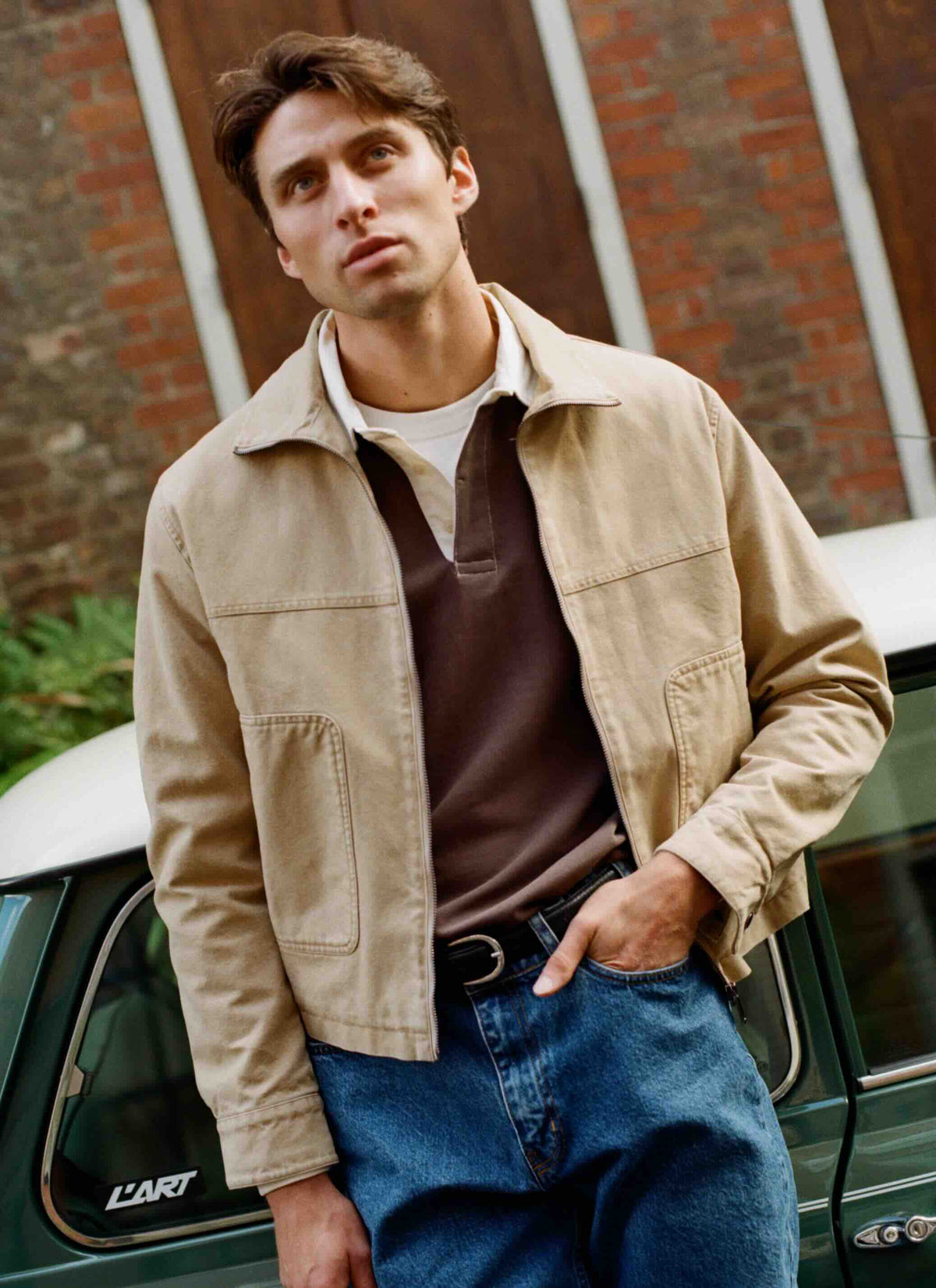 A model wearing the Percival brown Editor embroidered rugby polo shirt layered under a beige jacket outdoors.