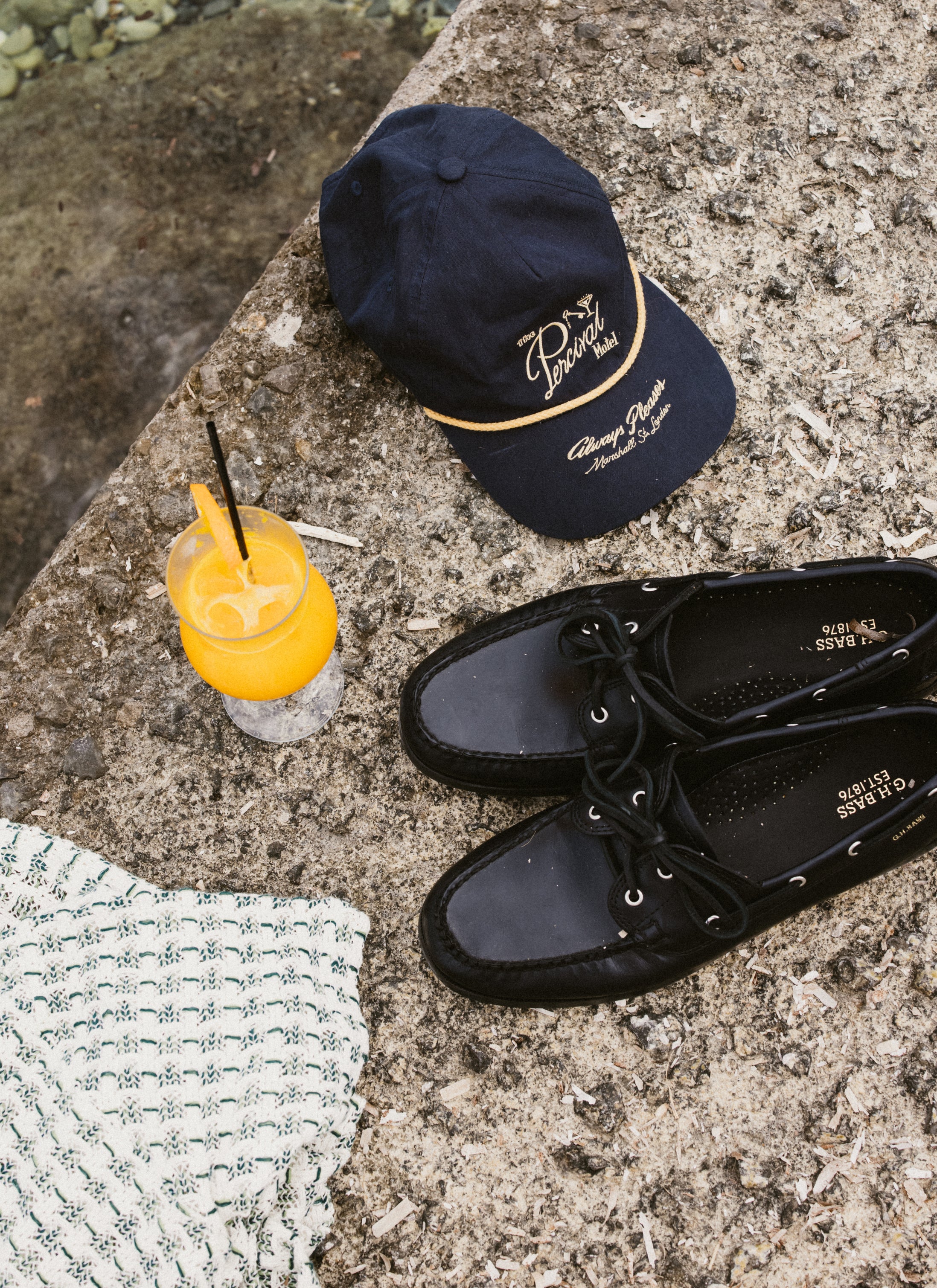 Navy Percival Kingsley soft trucker cap with gold embroidery, styled on a stone ledge with boat shoes and a summer drink.
