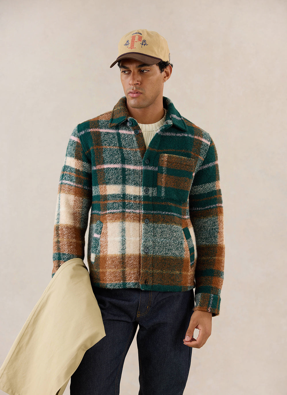 A model wears the multi-check wool Knowles Overshirt from Percival with a patterned cap and dark denim jeans.