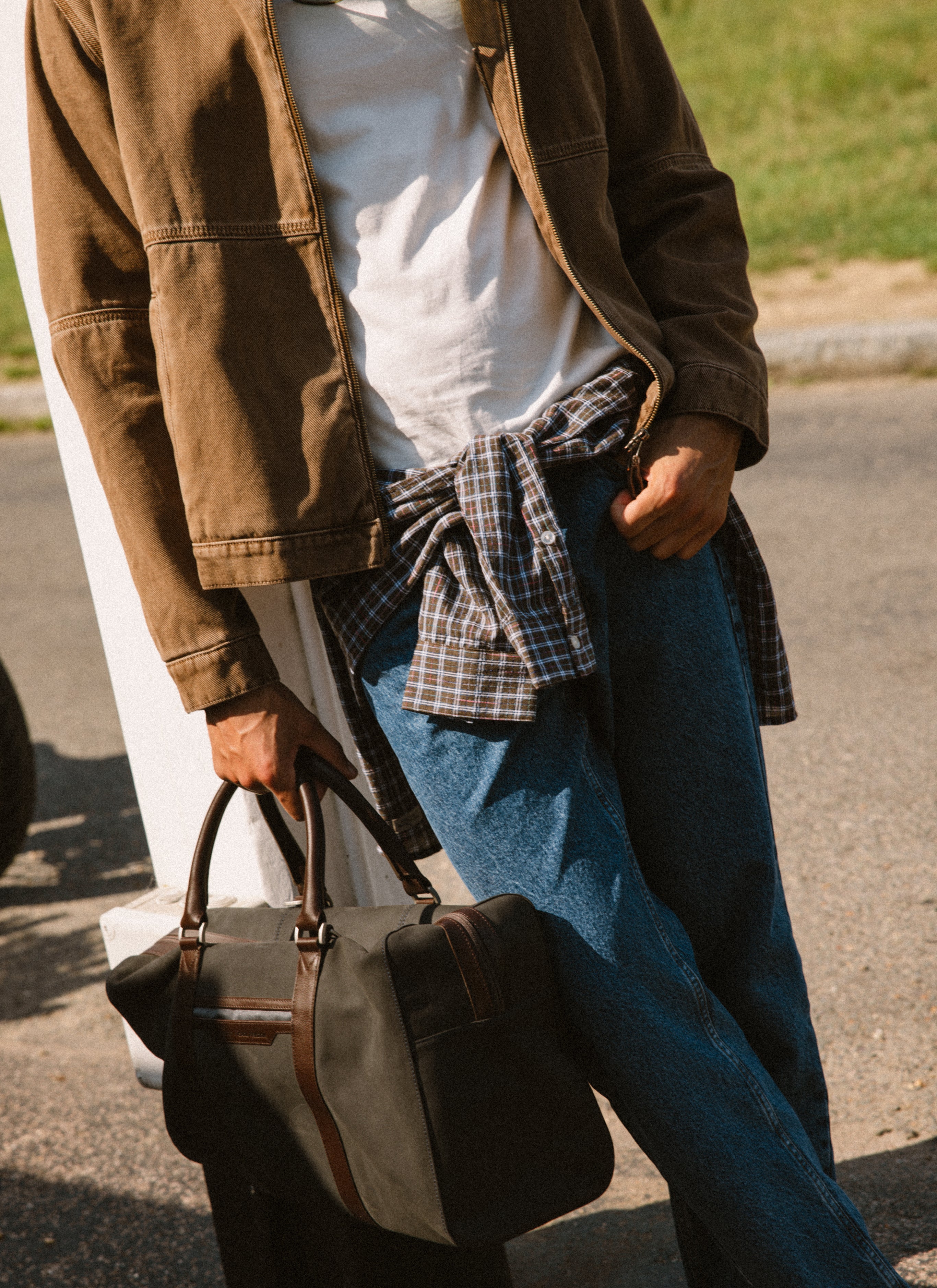 A model wears the Percival mocha cotton twill jacket, styled with a white tee, blue jeans, and a dark holdall.