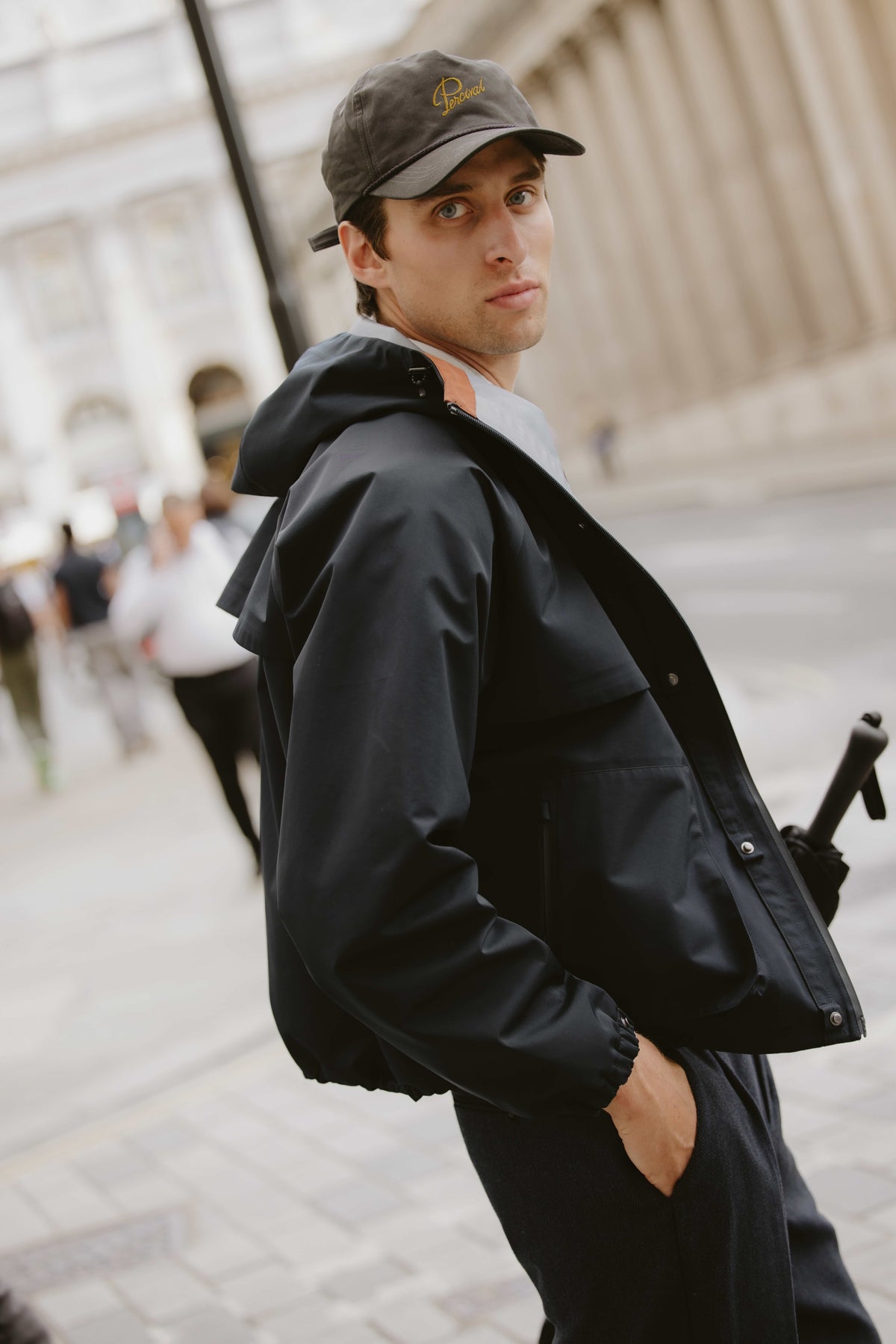 A model wearing the Percival navy blue Hanson waterproof rain jacket and a cap while standing outdoors on an urban street.