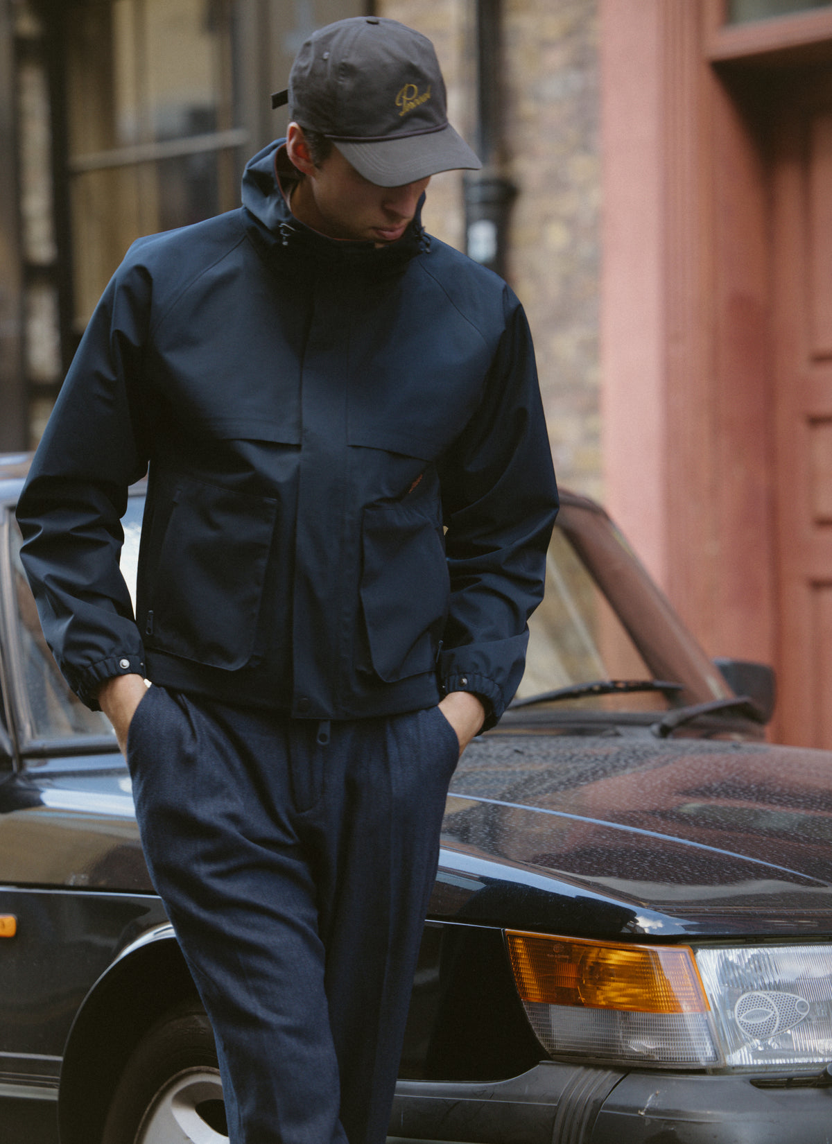 A model wearing the Percival navy blue Hanson waterproof rain jacket and a cap while standing outdoors near a car.