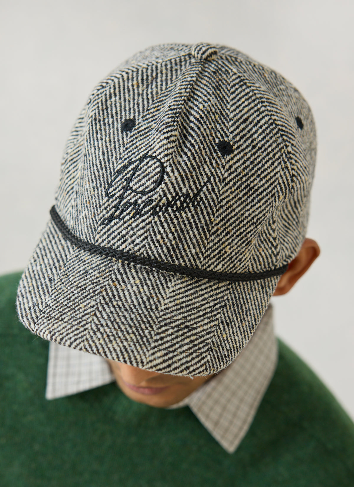 A model wearing the grey herringbone wool Percival soft trucker cap, featuring black embroidery and a rope trim.