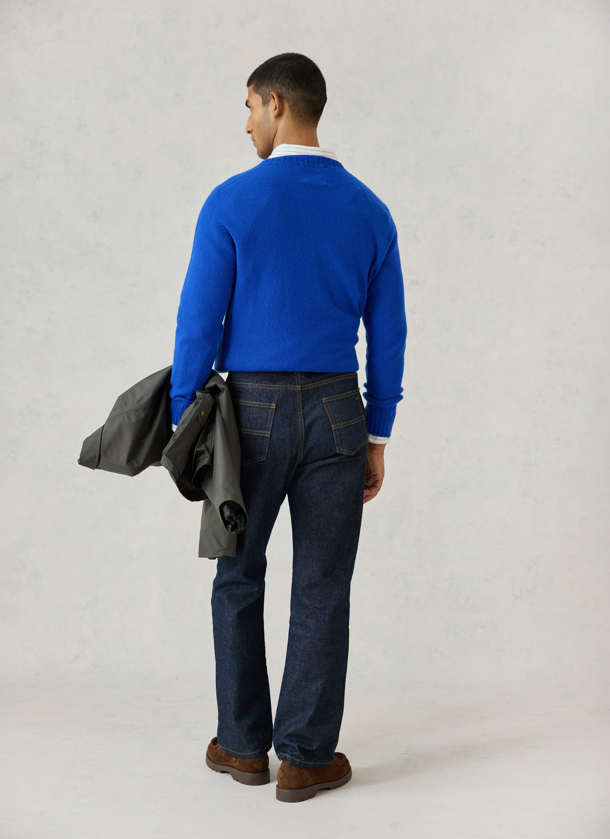 A model from the back wearing Percival's cobalt blue lambswool jumper and dark jeans, holding a green jacket.