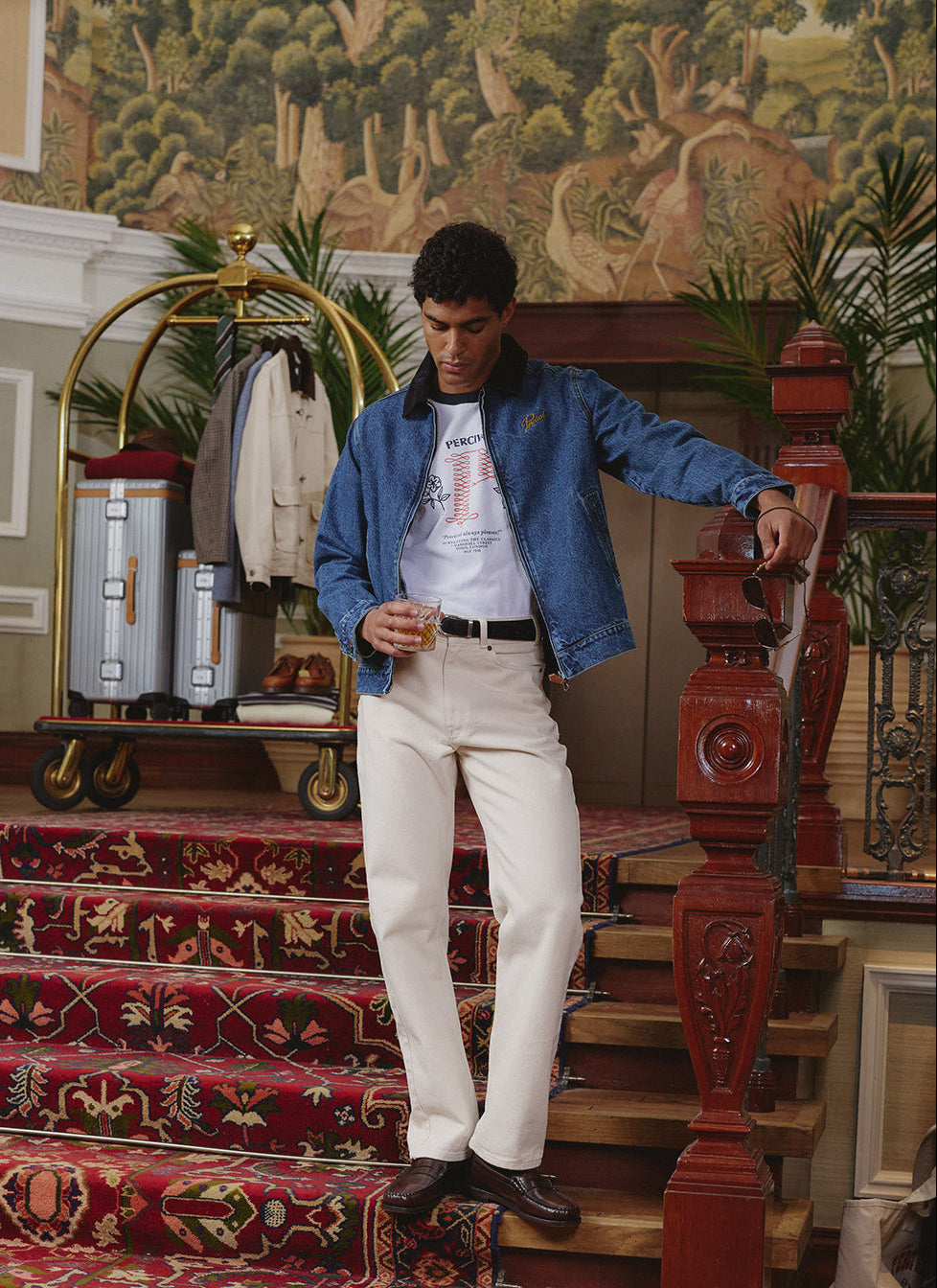 A model wearing a Percival denim jacket over a white ringer t-shirt and cream jeans, holding a glass on stairs.