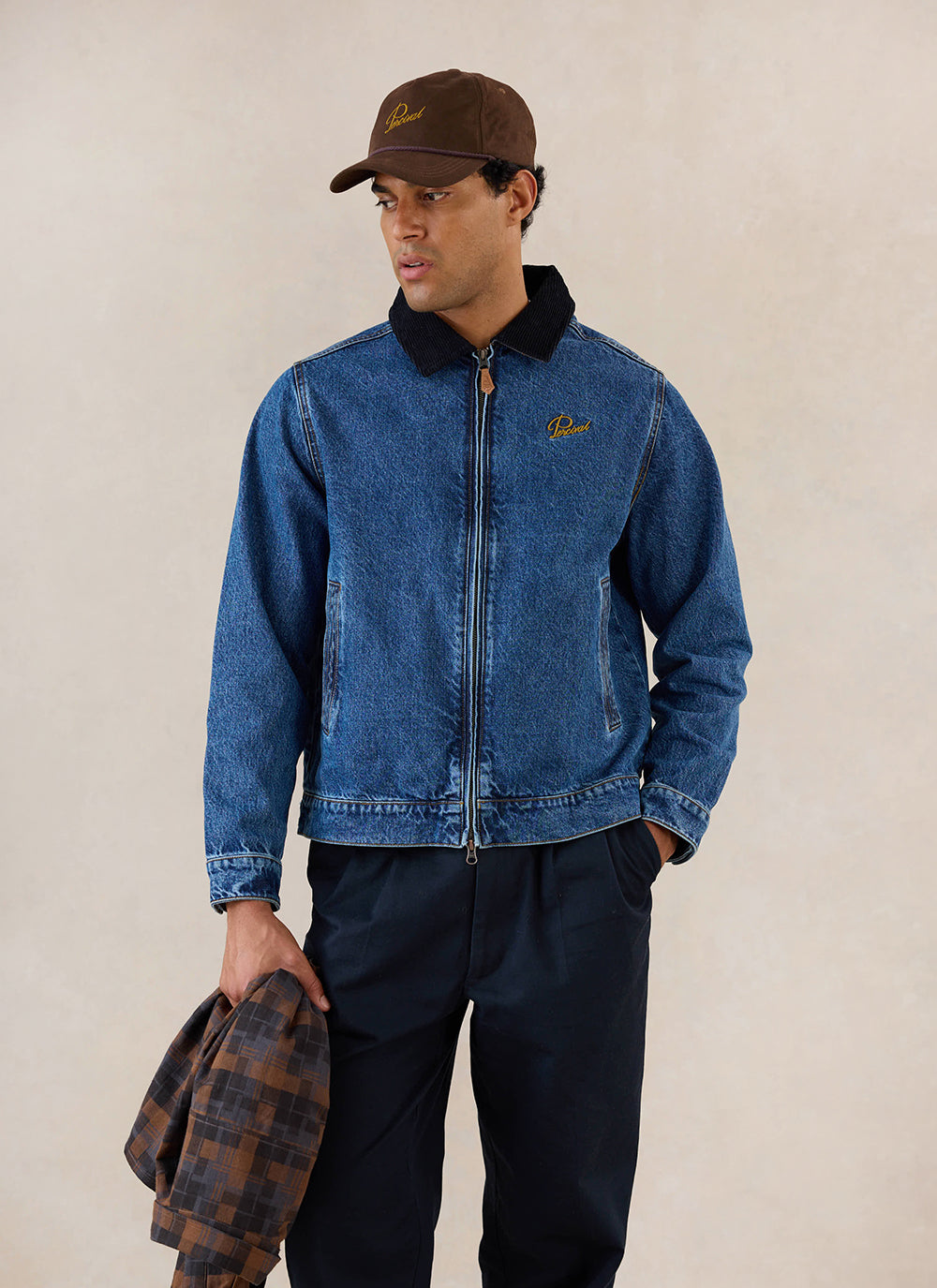 A model wears the mid wash blue Percival Mackey Zip Jacket in denim, styled with dark trousers and a brown cap.