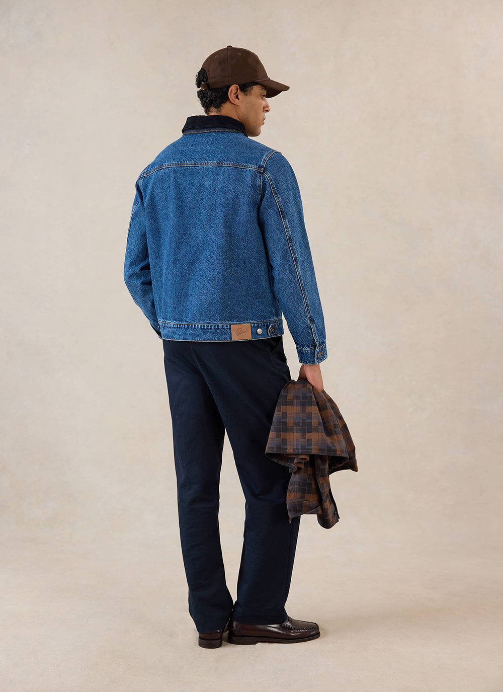 A model from behind wears the mid wash blue Percival Mackey zip jacket, paired with dark trousers and a brown cap.