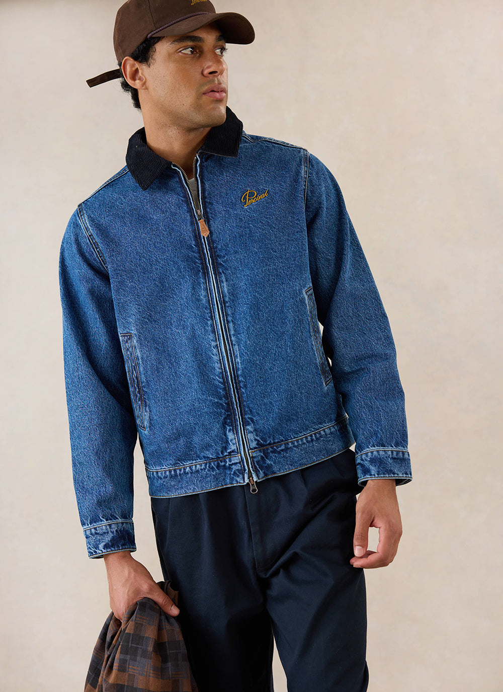 A model wears the mid wash blue Percival Mackey zip jacket featuring a contrast collar, styled with dark trousers and a cap.