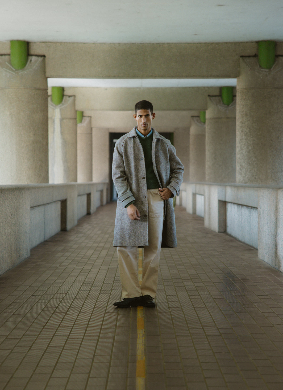 A model wearing the Percival grey wool herringbone car coat in a concrete walkway with large columns.