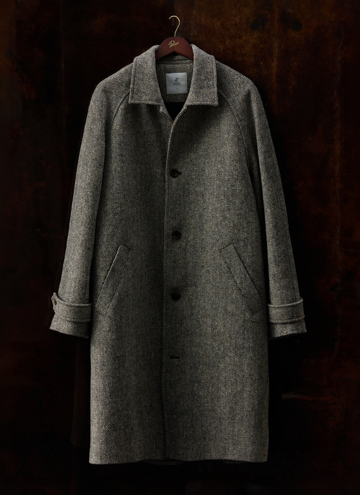 Grey herringbone wool car coat by Percival hanging on a wooden hanger against a dark textured background.