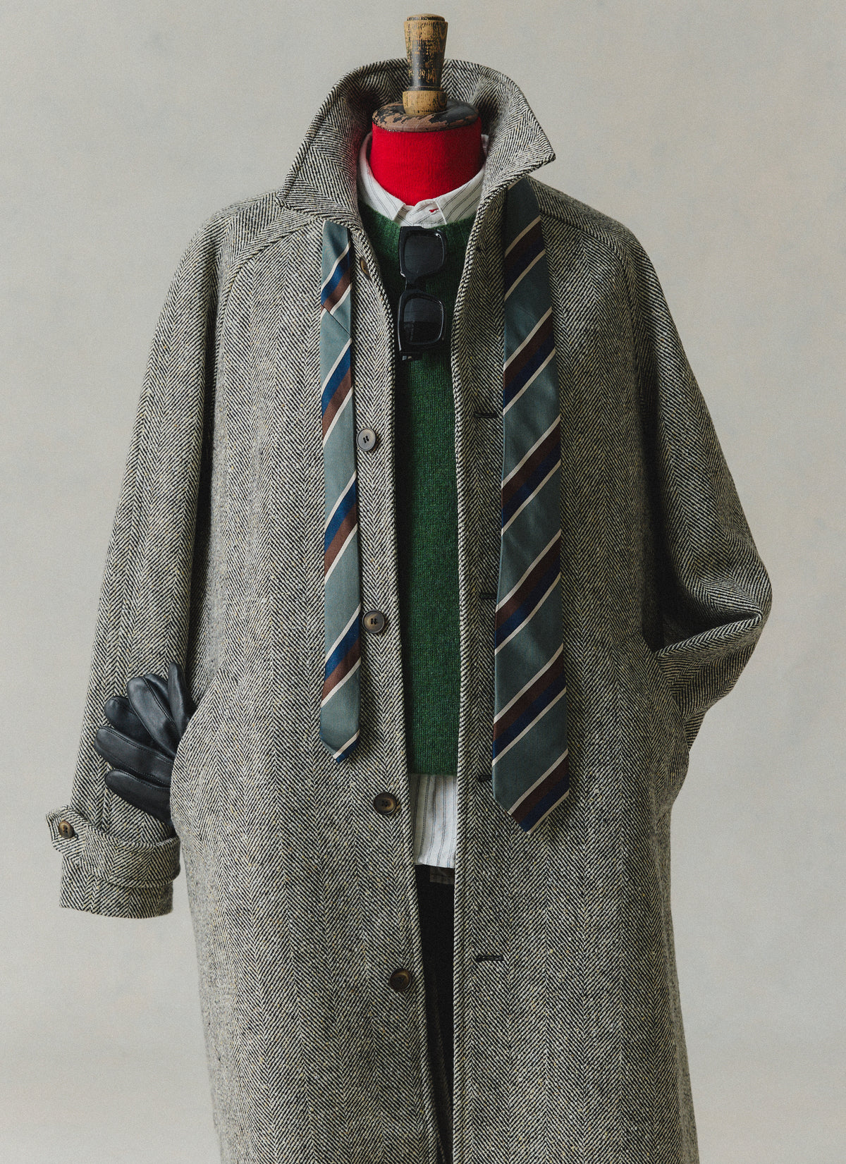 A herringbone wool car coat styled over a green lambswool jumper and striped tie on a mannequin.