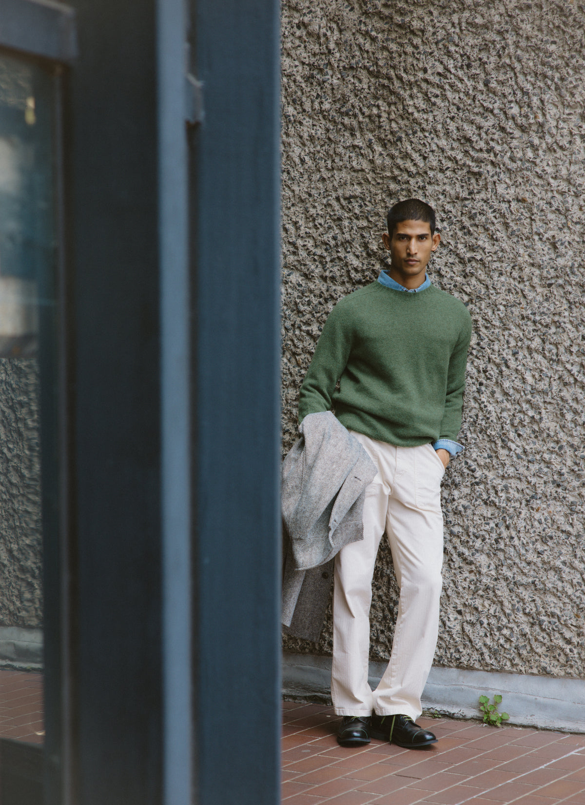 A model wearing the Percival forest green lambswool jumper and light trousers, holding a coat against a textured wall.