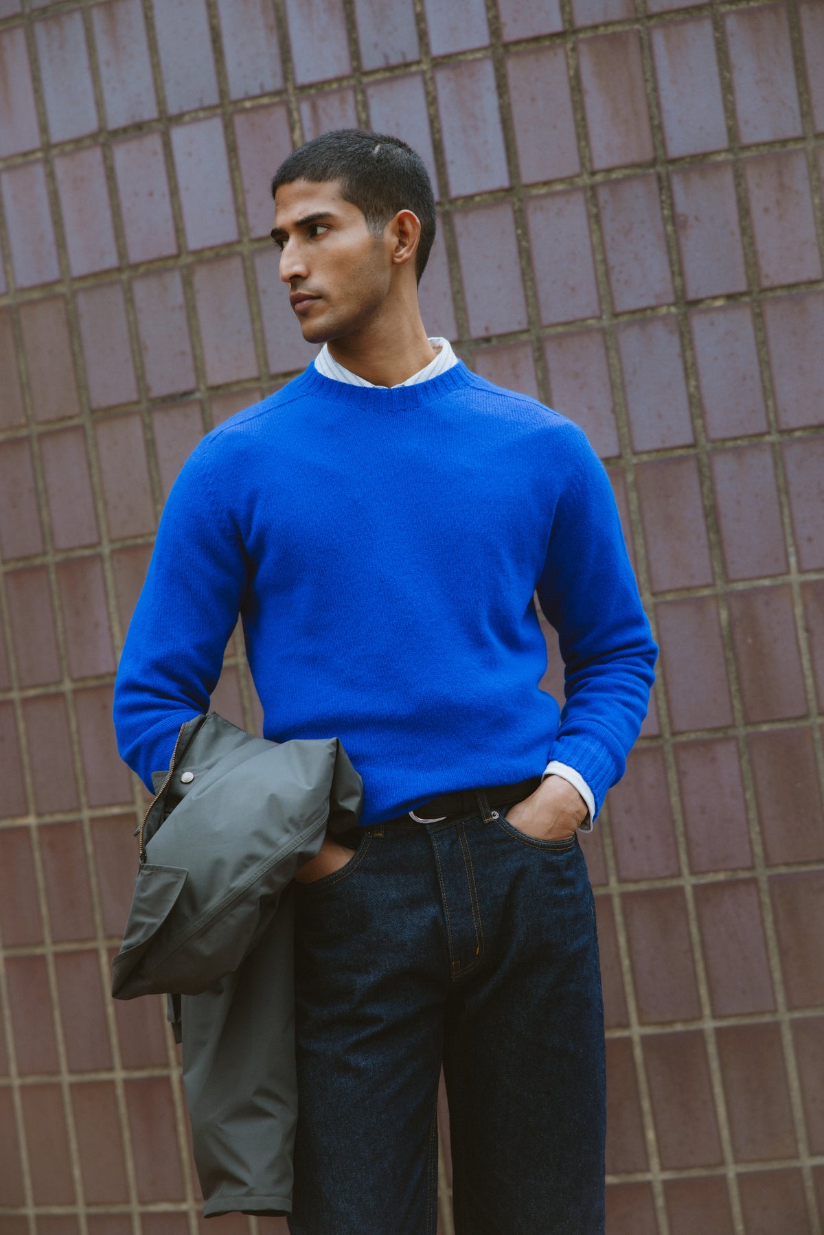 A model wearing Percival's cobalt blue lambswool jumper with a white shirt and dark jeans, holding a jacket.