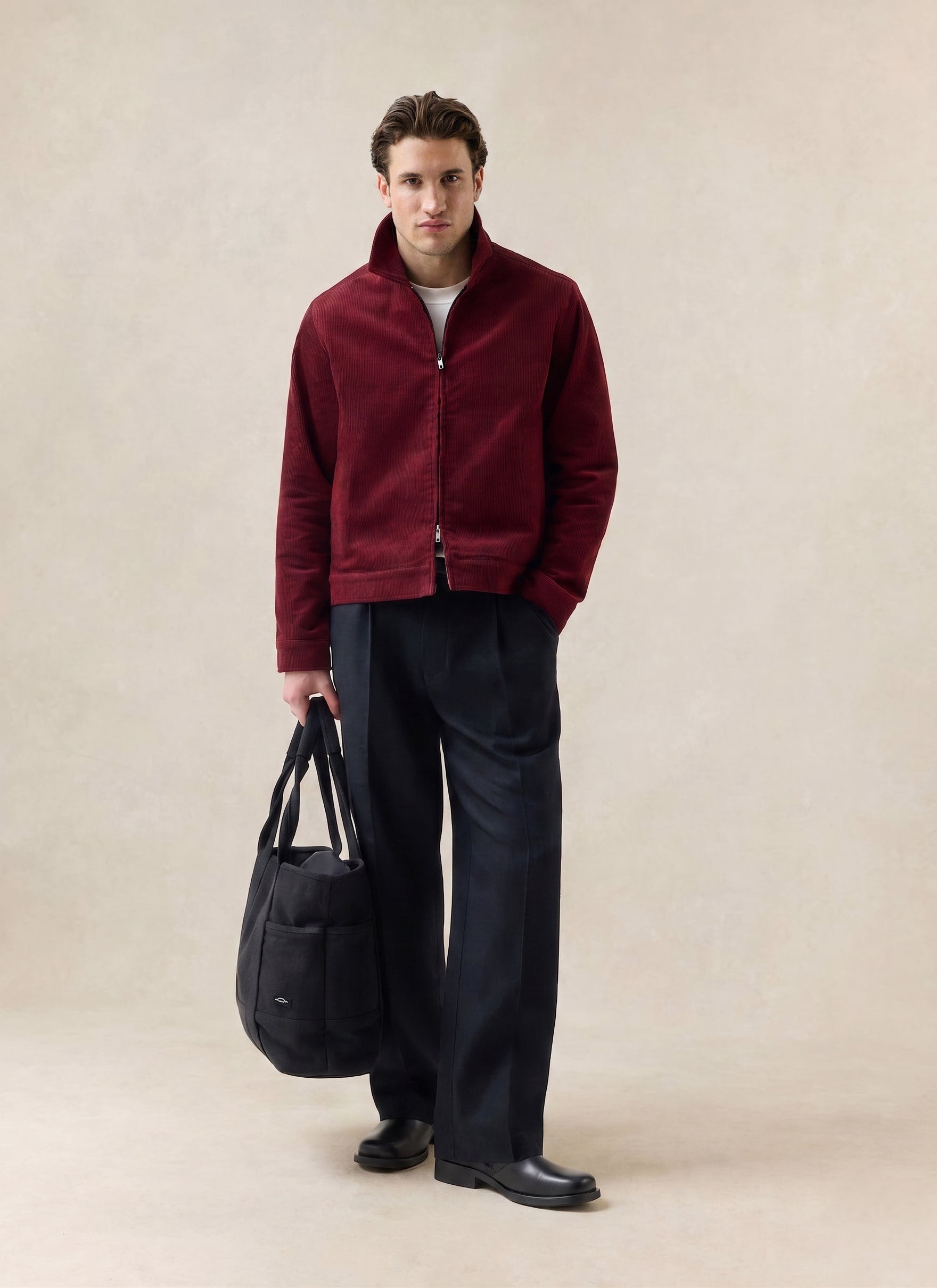 A model wears the burgundy Percival cord jacket, styled with dark trousers and holding a black tote bag in a studio.