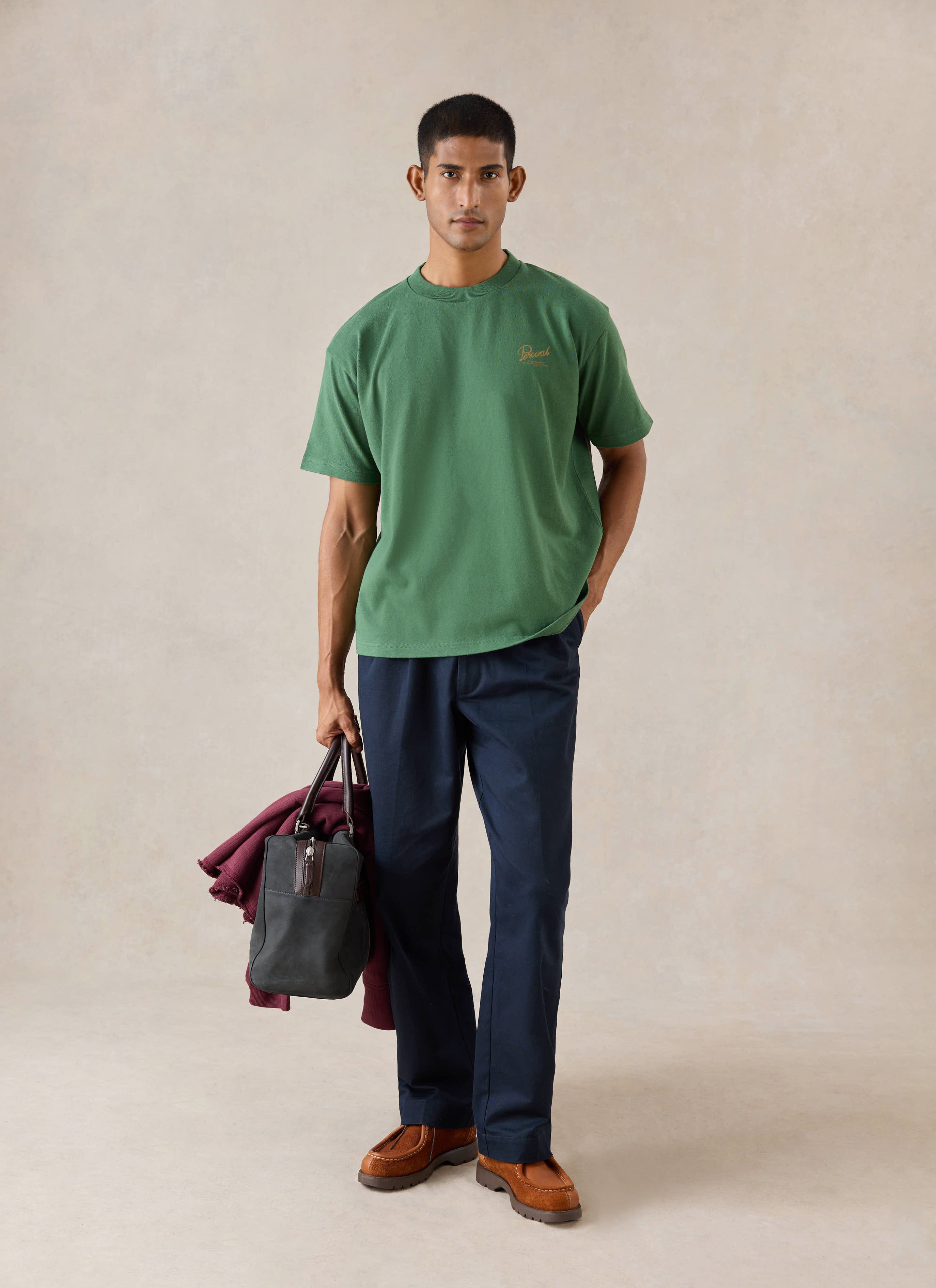 A model wears the forest green Percival Marley boxy t-shirt with navy trousers, holding a bag and a burgundy jacket.