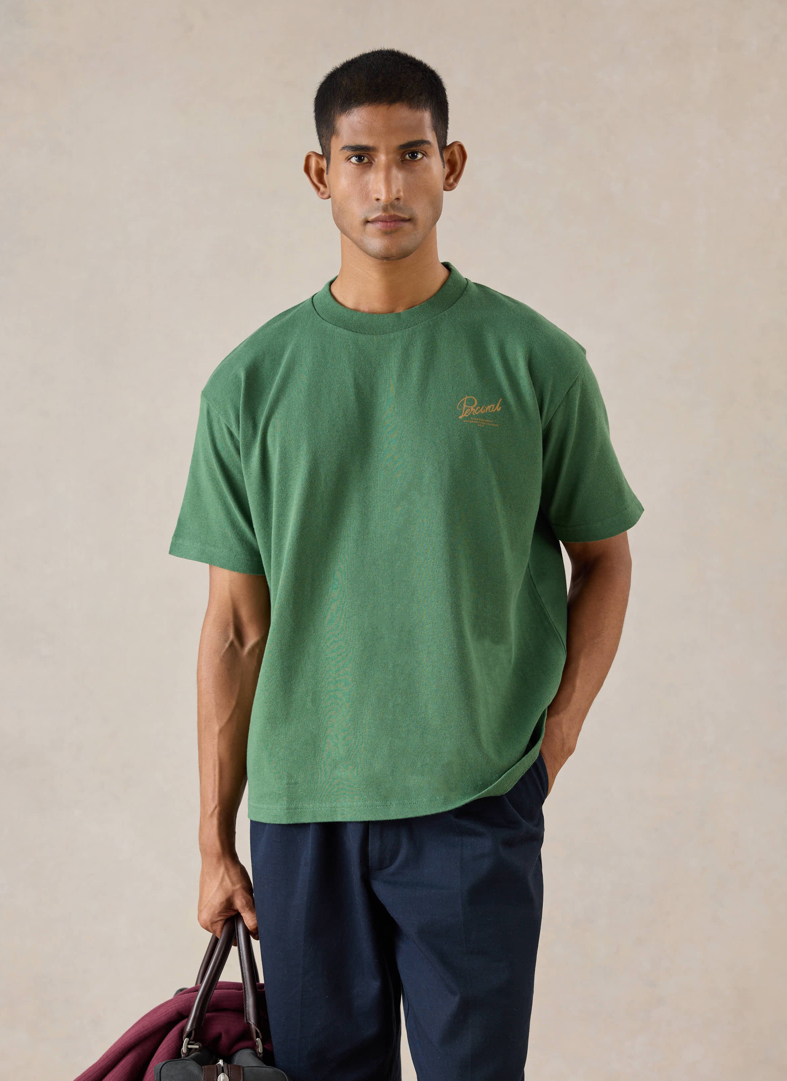 A model wears the forest green Percival Marley boxy t-shirt in organic cotton, styled with dark trousers.
