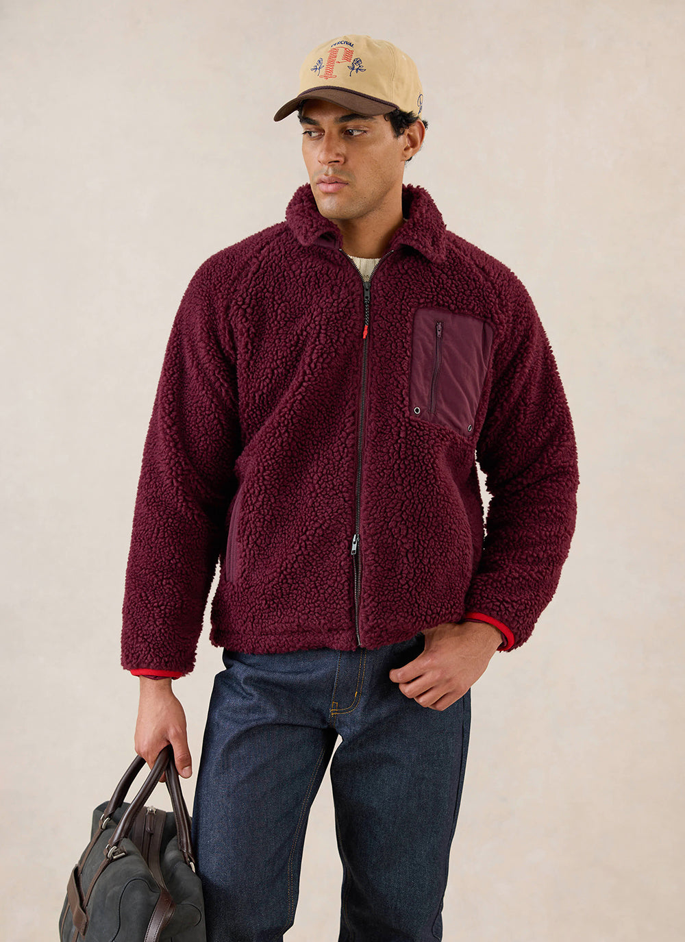 A model wears the burgundy Percival fleece jacket with a cap and bag in a studio shot.