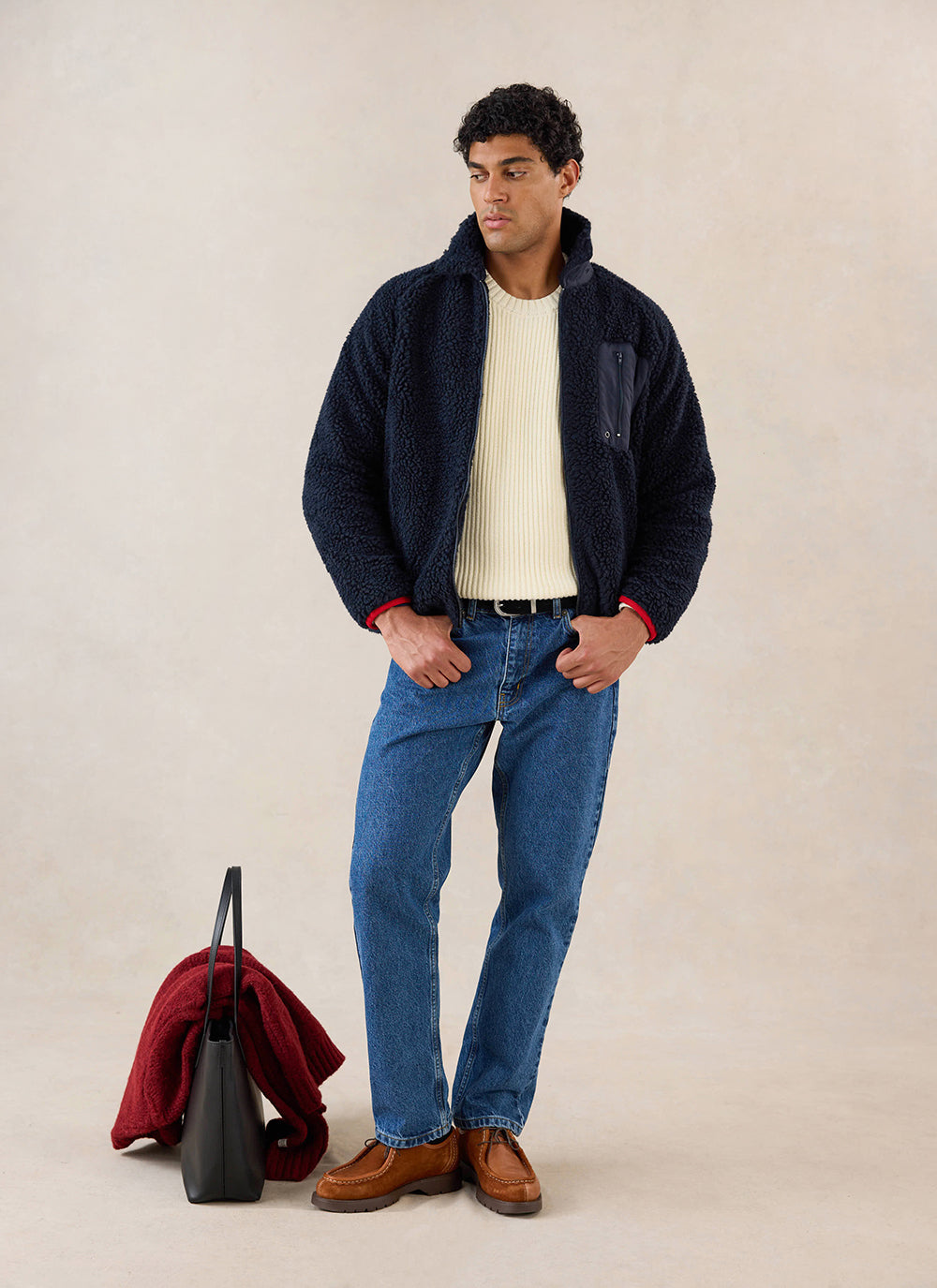 A model wears the navy wool fleece jacket from Percival over a cream sweater, with blue jeans and brown shoes in a studio.