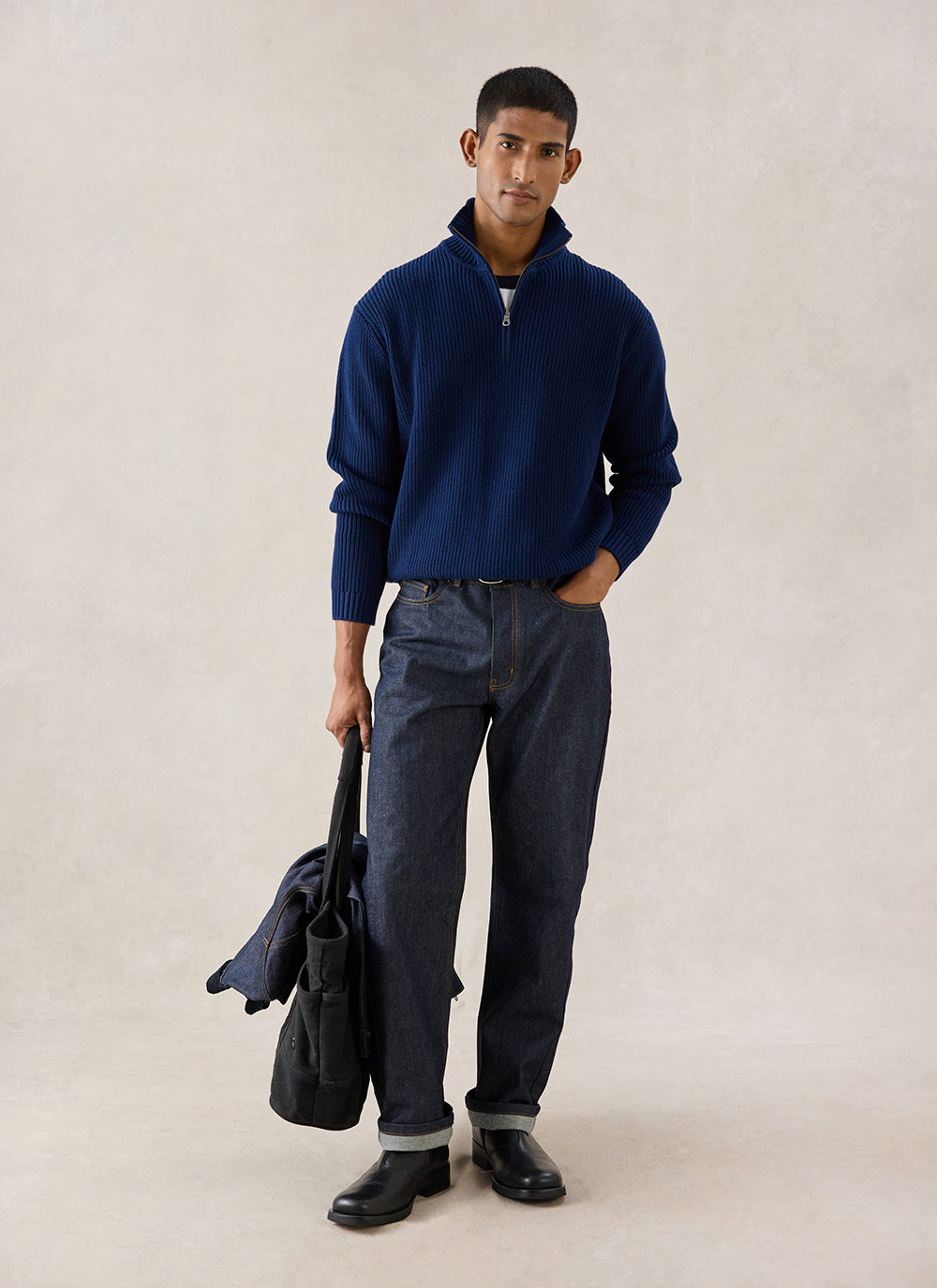 A model wears the navy Meyer Boxy Quarter Zip from Percival with dark jeans, holding a bag in a studio.