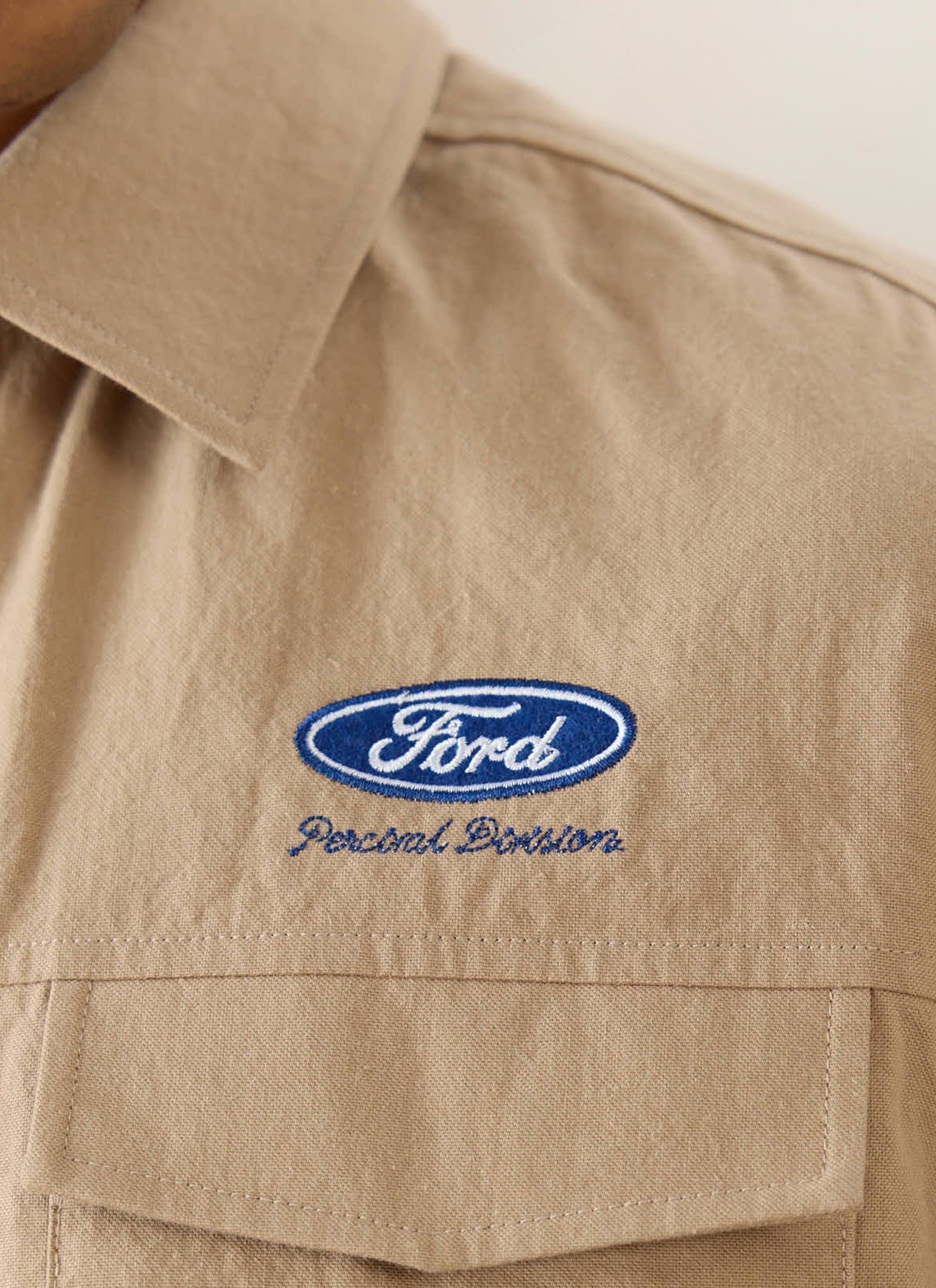 Close-up of the blue Ford logo and Percival Division embroidery on the beige cotton-hemp Miles Garage shirt.