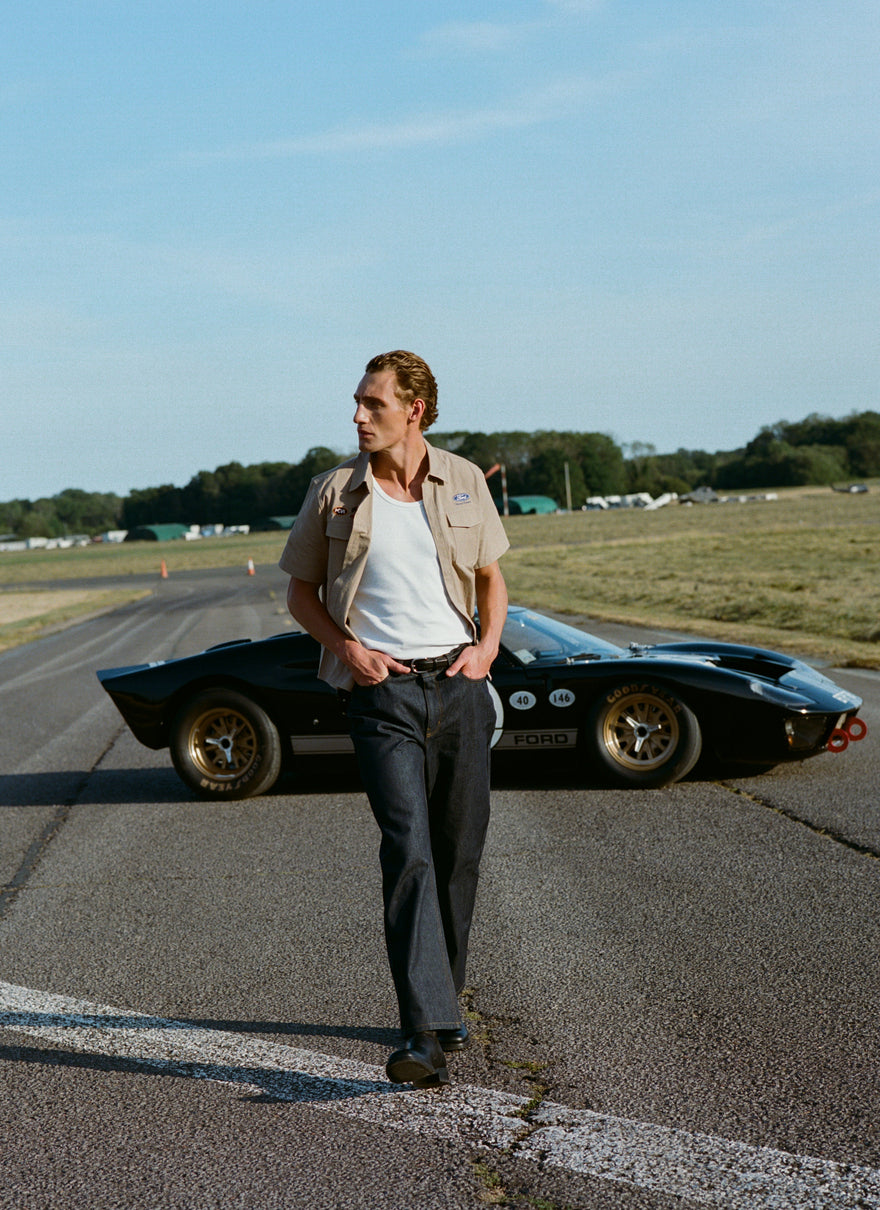 A model wears the beige Percival x Ford Miles Garage Shirt and dark heritage denim, posed by a vintage car on a race track.