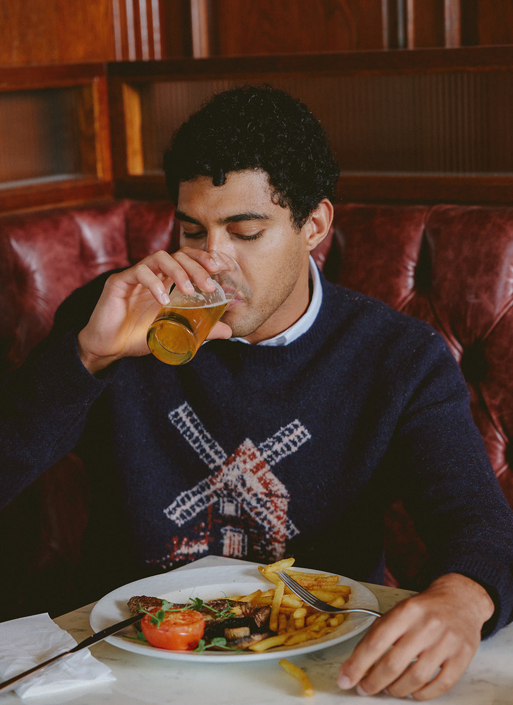 A model wearing the navy Percival Miller Jacquard Jumper with a windmill pattern, drinking a beer in a restaurant booth.
