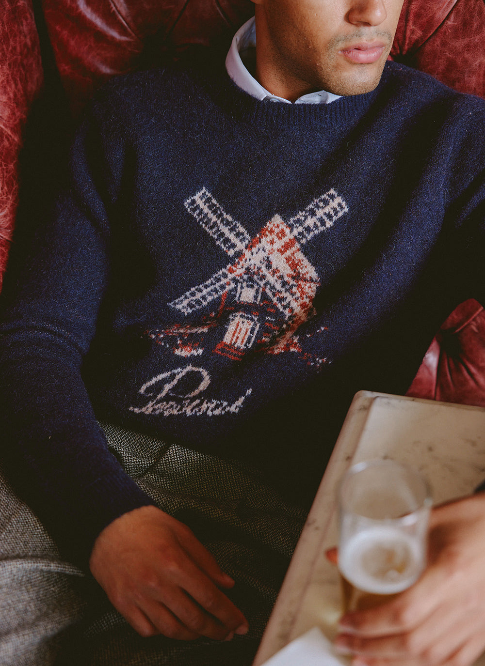 A model wearing the navy Percival Miller Jacquard jumper featuring a windmill graphic and script branding, seated indoors.