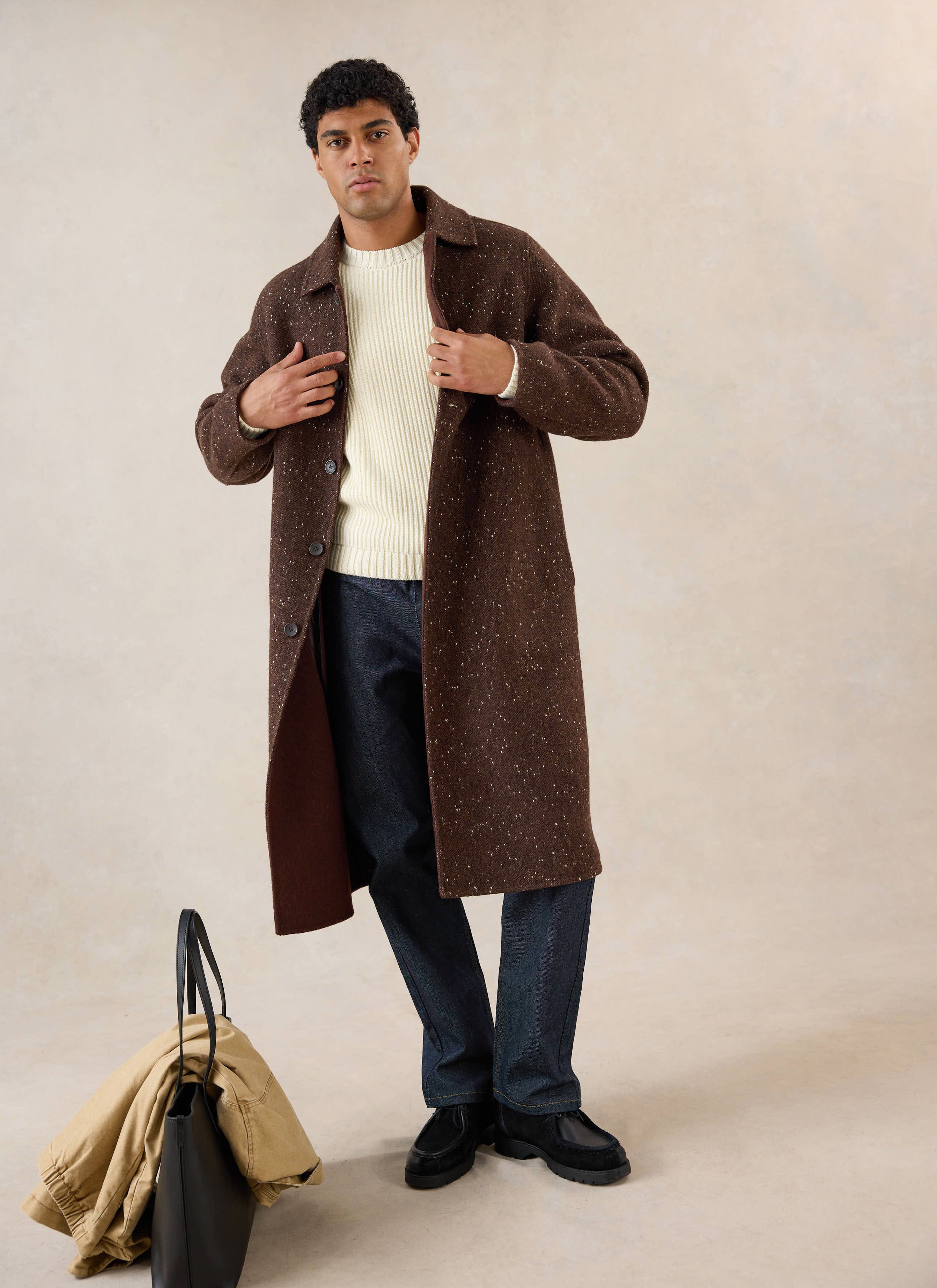A model wears the brown fleck Percival Minnesota overcoat over a cream knit jumper and dark trousers in a studio shot.