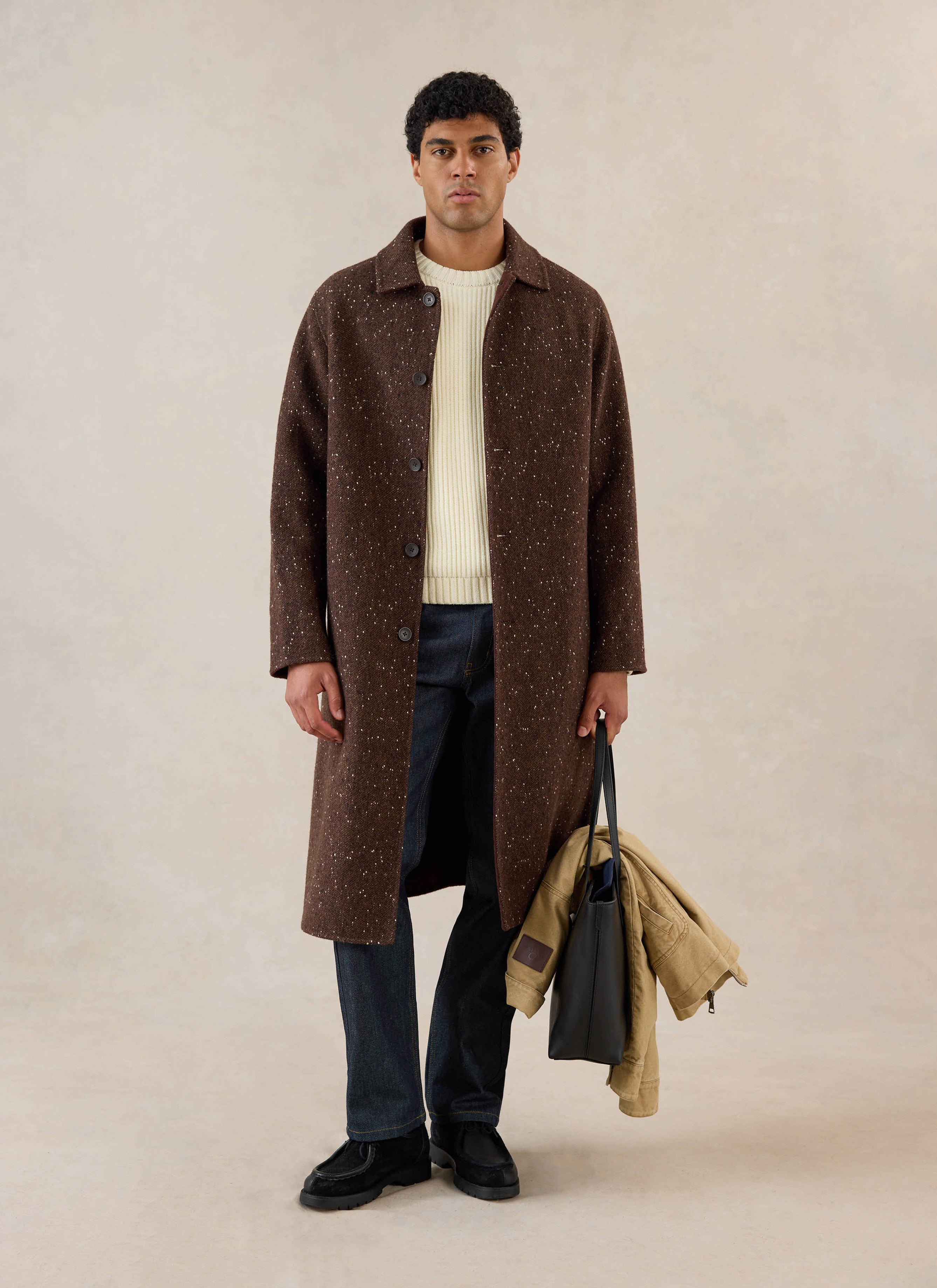 A model wears the brown fleck Percival Minnesota Overcoat over a cream knit jumper and jeans, holding a black bag.