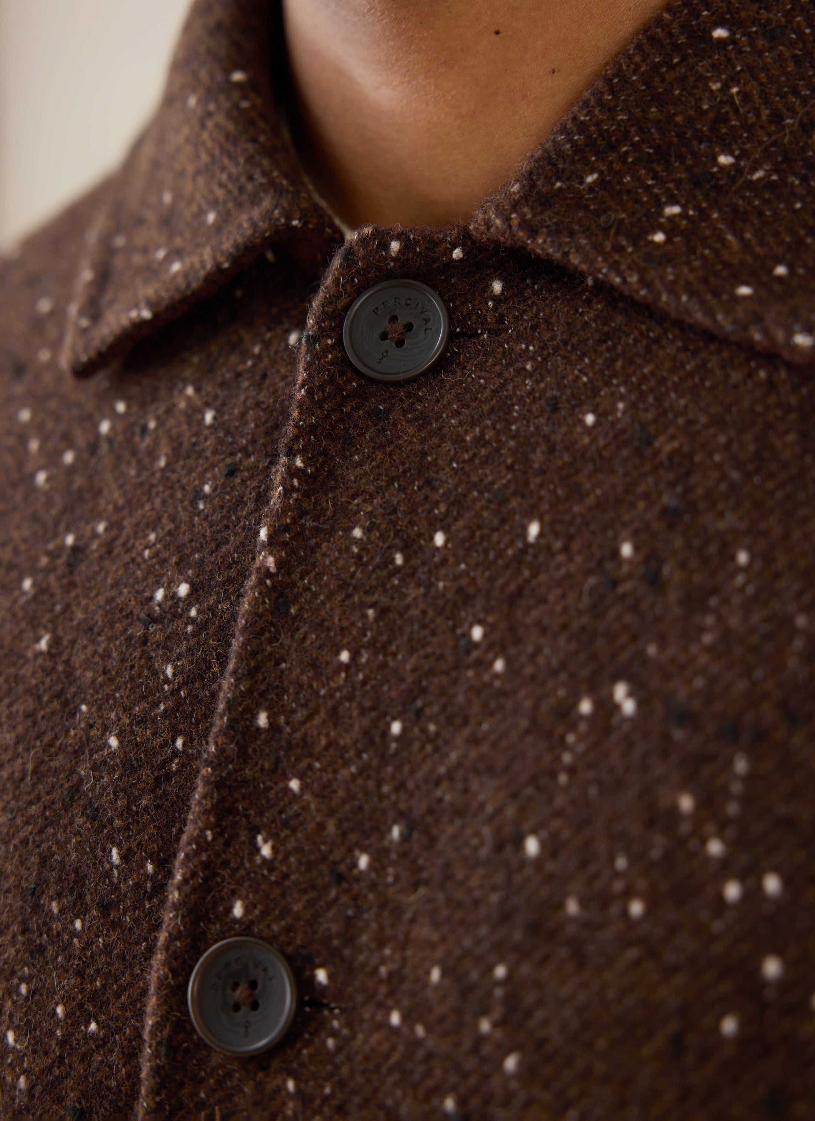 A close-up of the brown fleck twill Minnesota Overcoat from Percival, showing the textured fabric and branded button details.