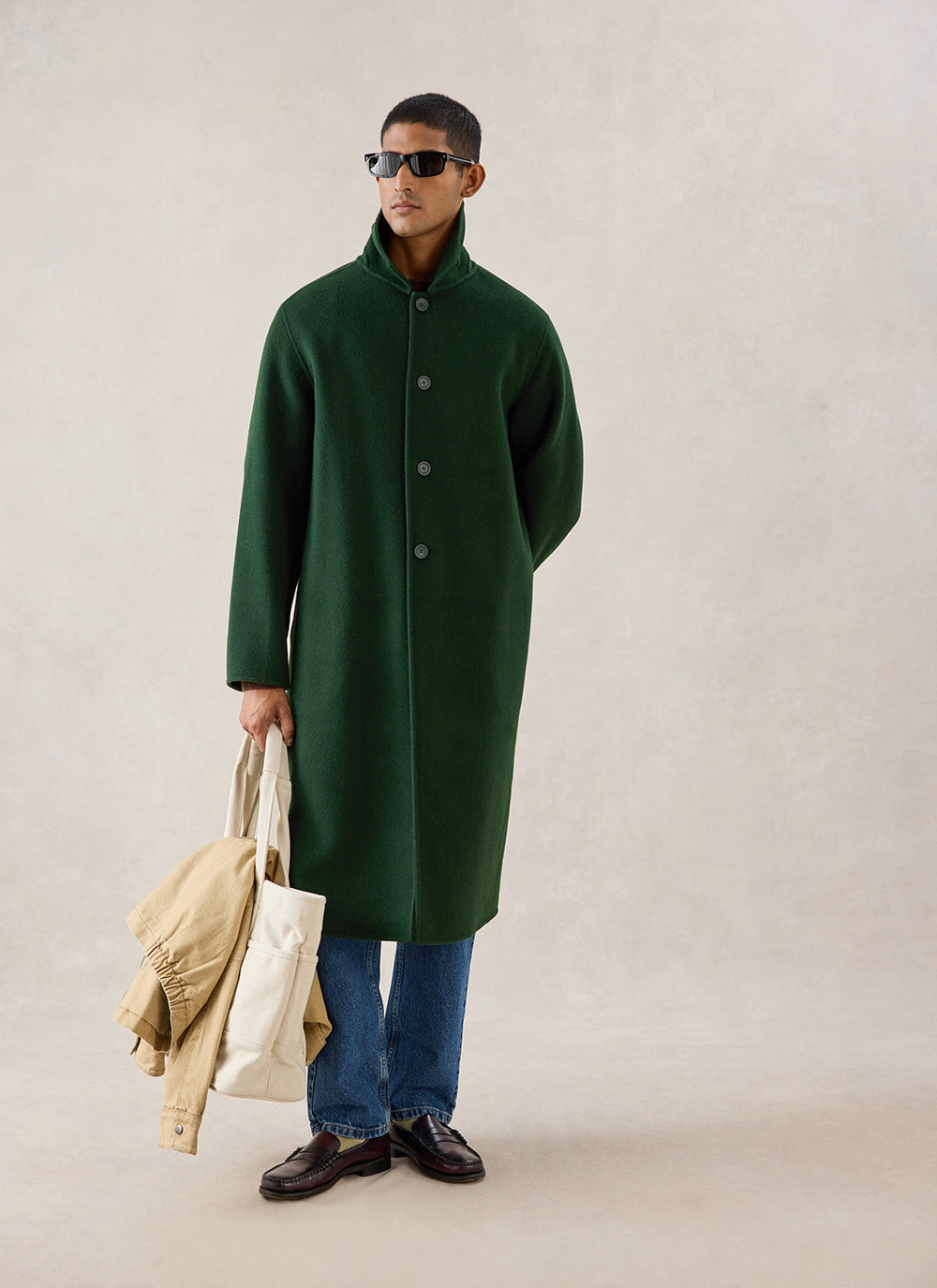 A model wears the forest green wool Minnesota Overcoat from Percival, paired with blue jeans, a tote bag and sunglasses.