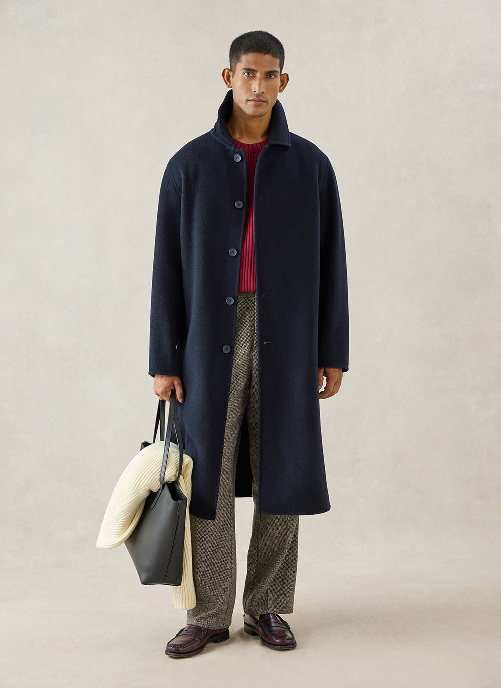 A model wears the navy wool Percival Minnesota Overcoat, styled with patterned trousers, a red jumper and a tote bag.