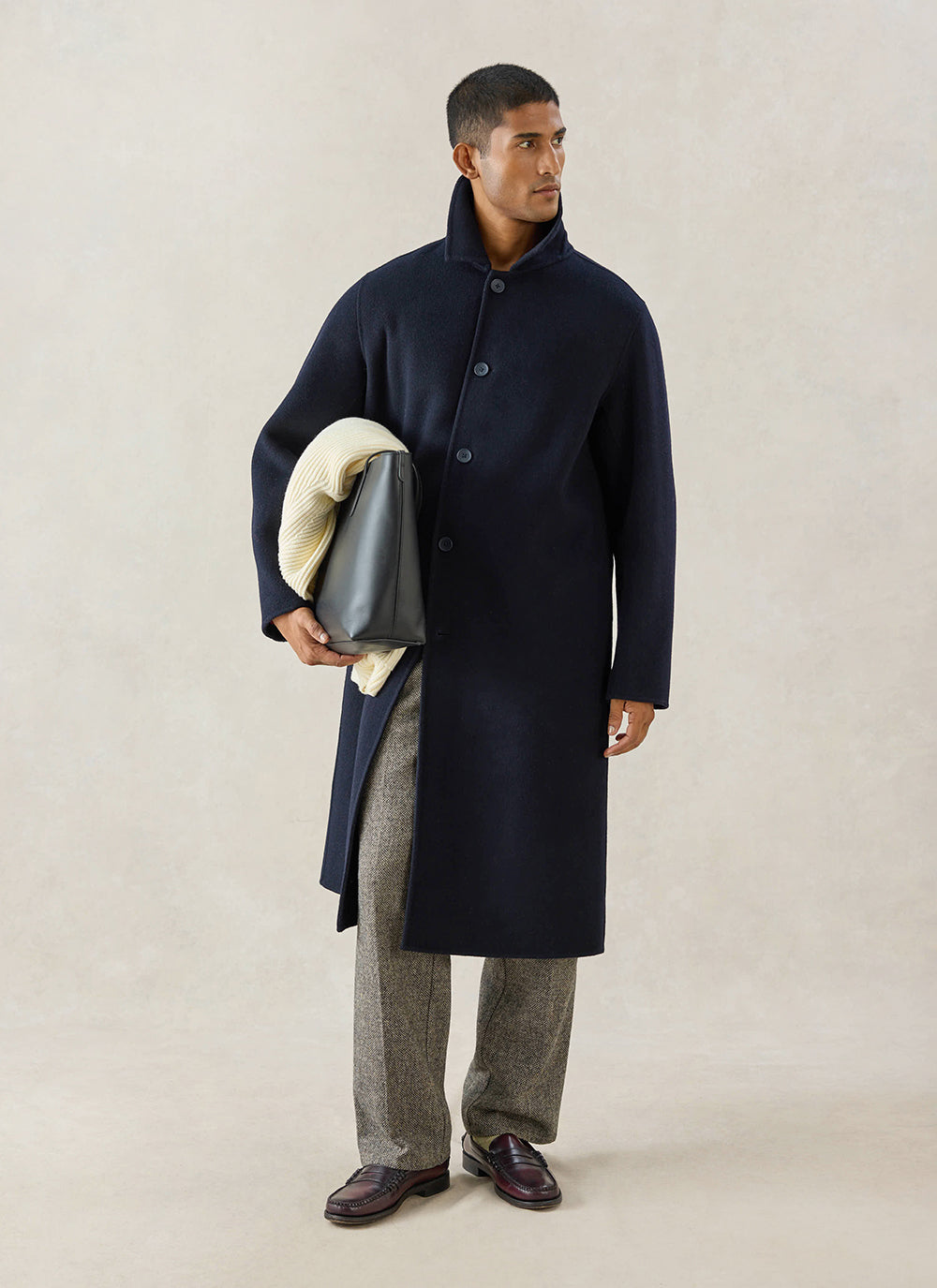 A model wears the navy wool Minnesota overcoat from Percival with tweed trousers, a tote bag and loafers in a studio.