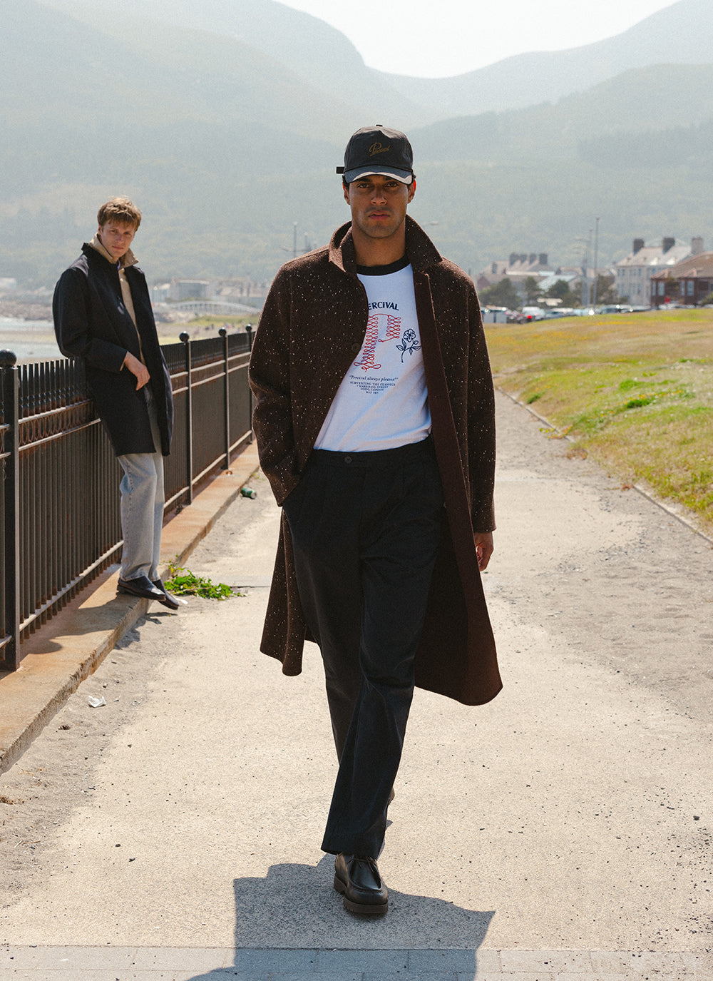 A model wearing a Percival brown fleck overcoat, charcoal cap, graphic t-shirt and dark trousers outdoors by the coast.