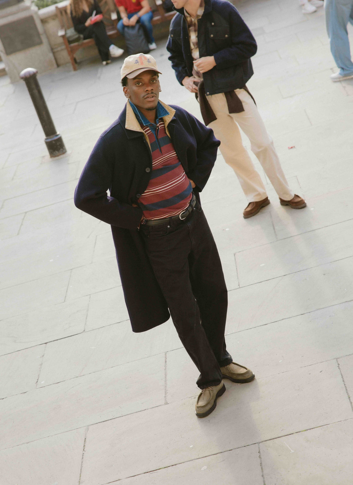 A model wearing a burgundy striped rugby shirt, denim collar, navy overcoat, and tan cap outdoors on a paved street.