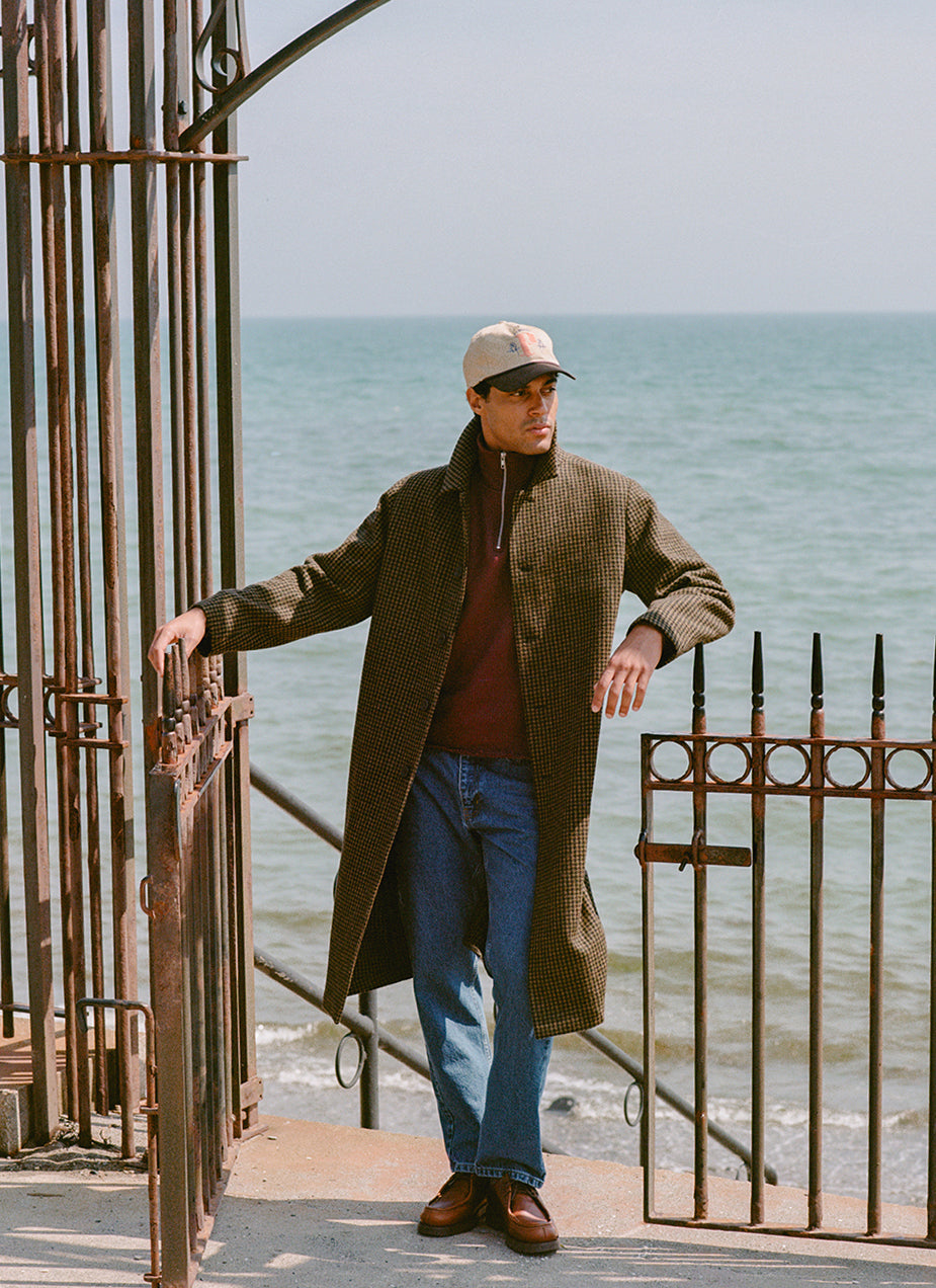 A model wears the Percival Minnesota wool overcoat in Watcher Check over a burgundy zip jumper and jeans.