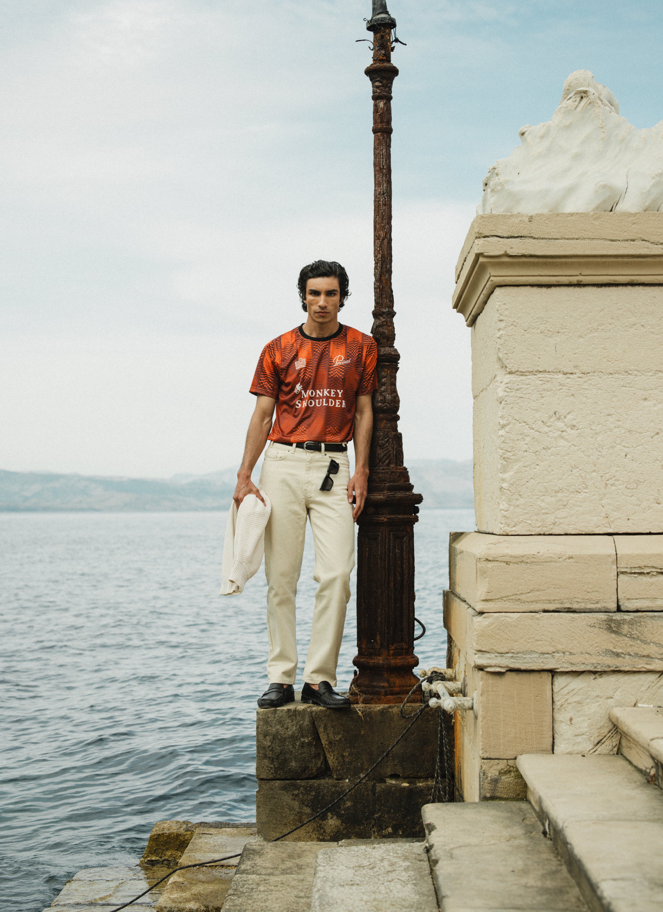 Percival x Monkey Shoulder orange football shirt with pixelated stripes, worn by a model standing by a coastal pier.