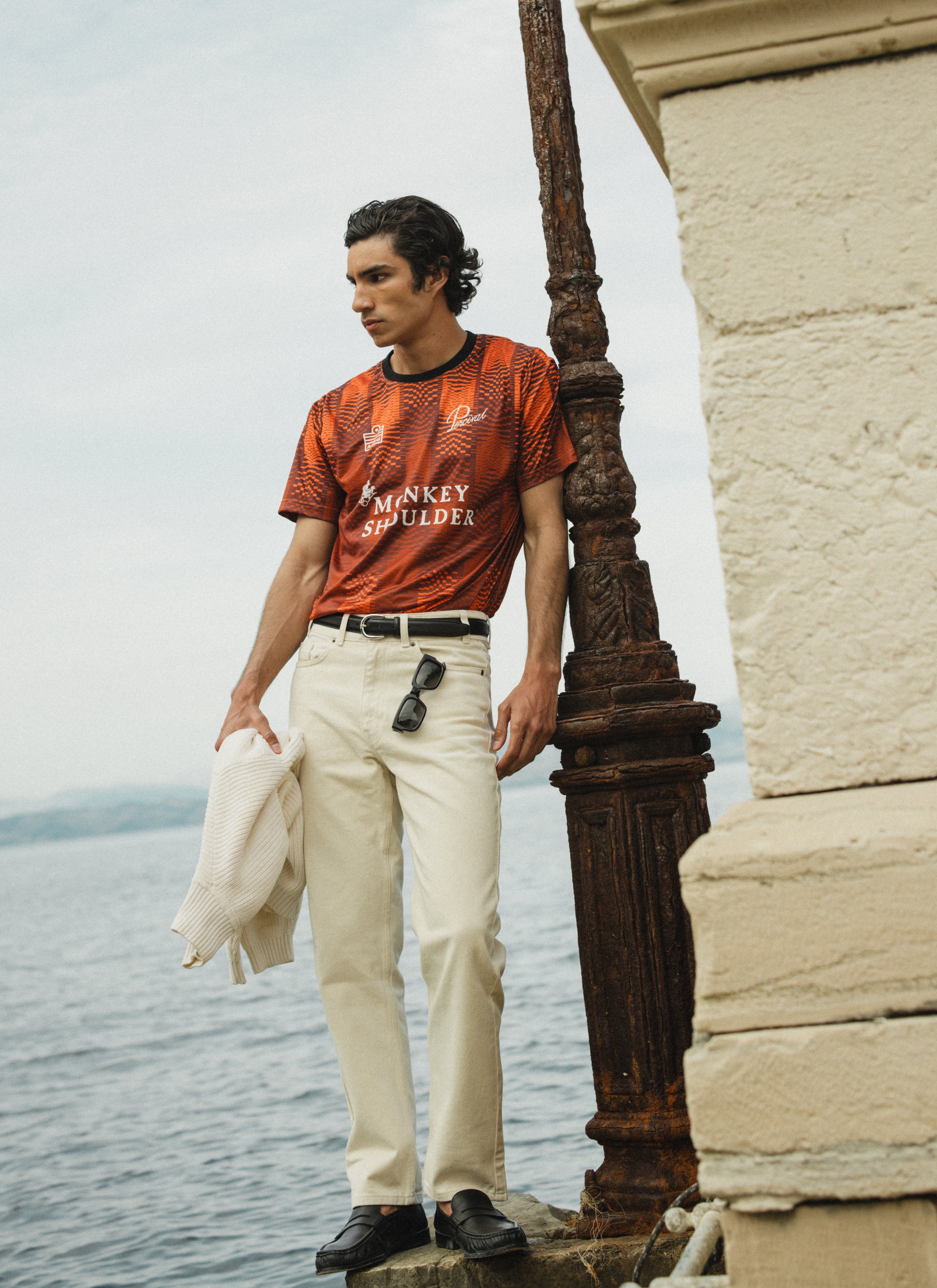 Percival x Monkey Shoulder limited-edition orange football shirt with pixel-distortion stripes, worn by a model by the sea.